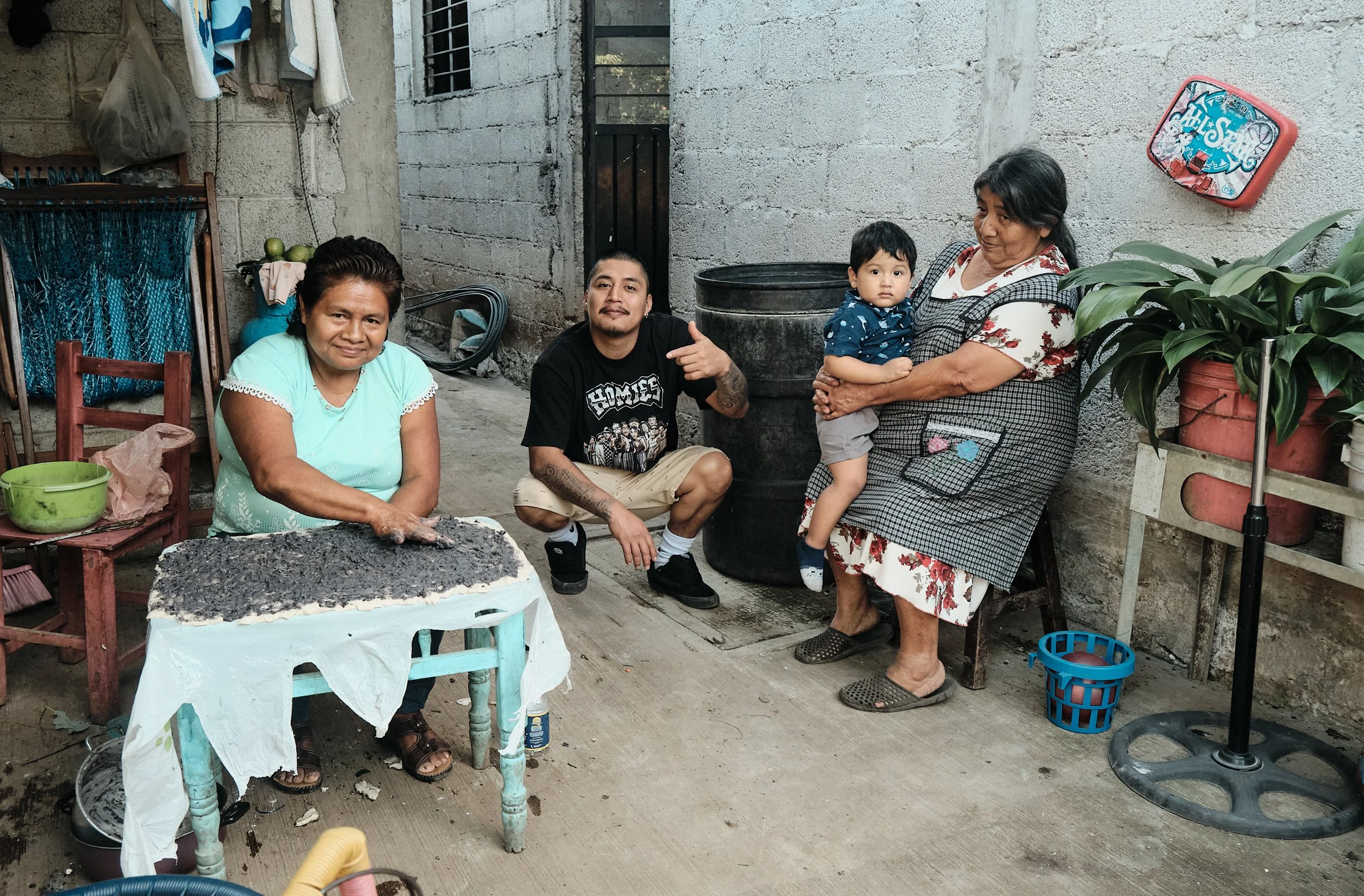Una familia de cinco personas en un espacio exterior, entre ellas un niño, probablemente abuela, madre, padre y un niño pequeño, en un entorno rústico con paredes de cemento. La abuela está sentada en una silla, la madre sostiene al niño en su regazo