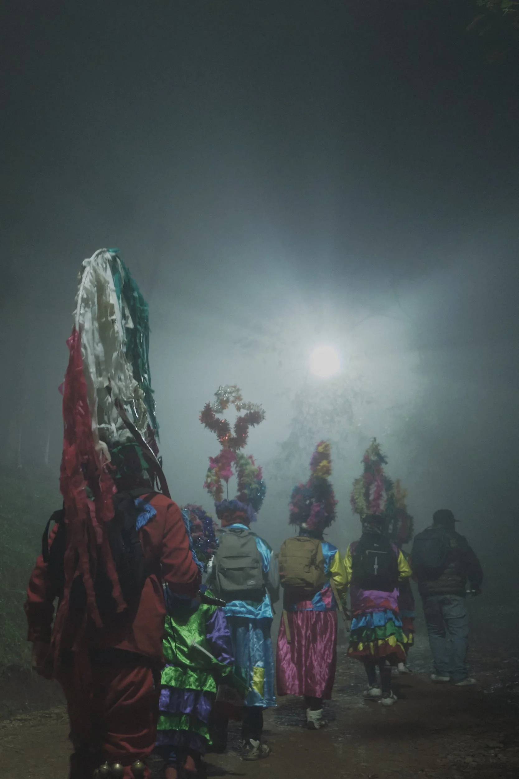 Grupo de personas participando en una ceremonia nocturna, con algunos llevando adornos coloridos en la cabeza, en un ambiente con poca iluminación y un cielo oscuro.