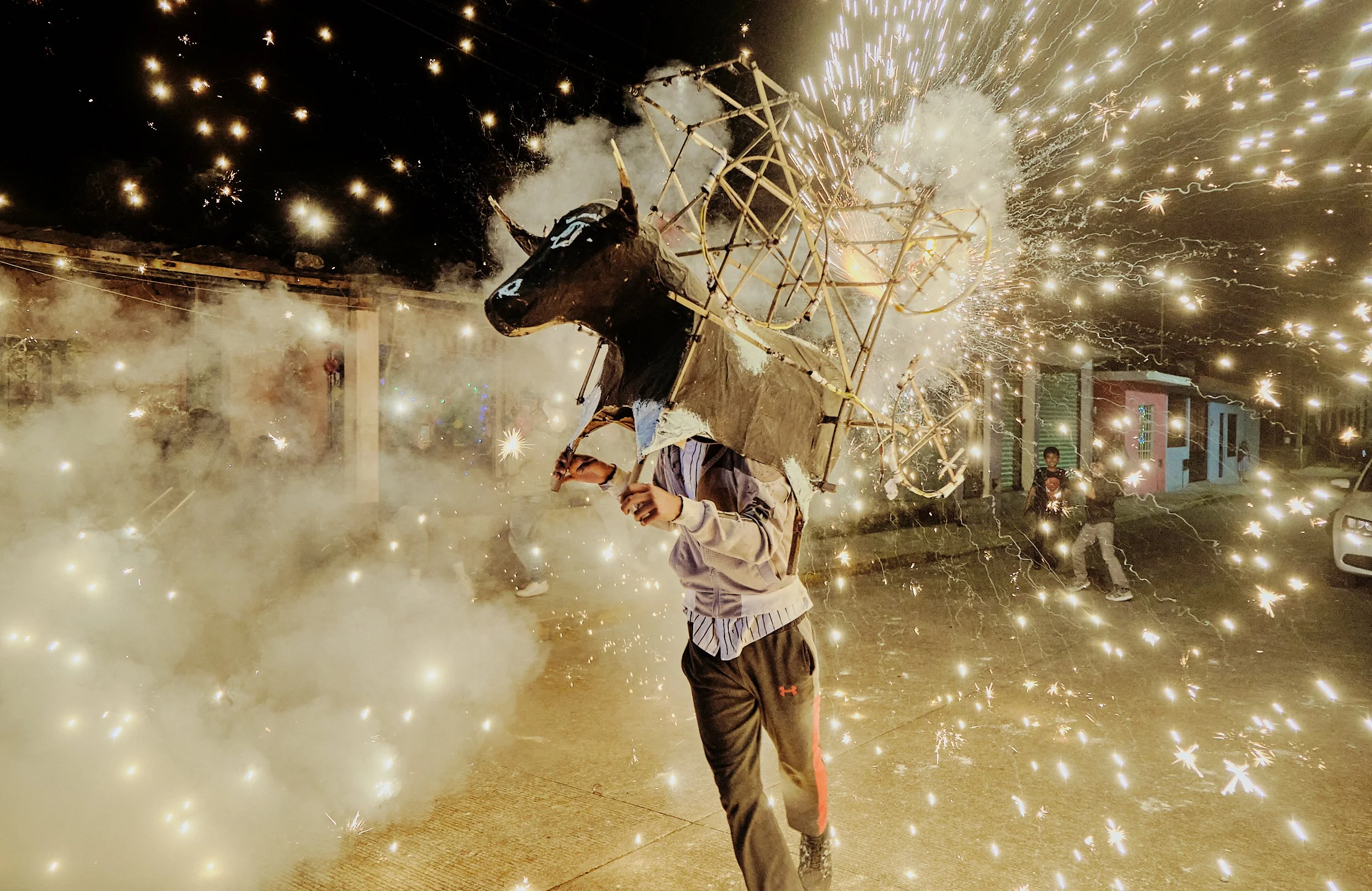 Persona sosteniendo una figura de madera o cartón en forma de toro en medio de fuegos artificiales y chispas durante la celebración nocturna.