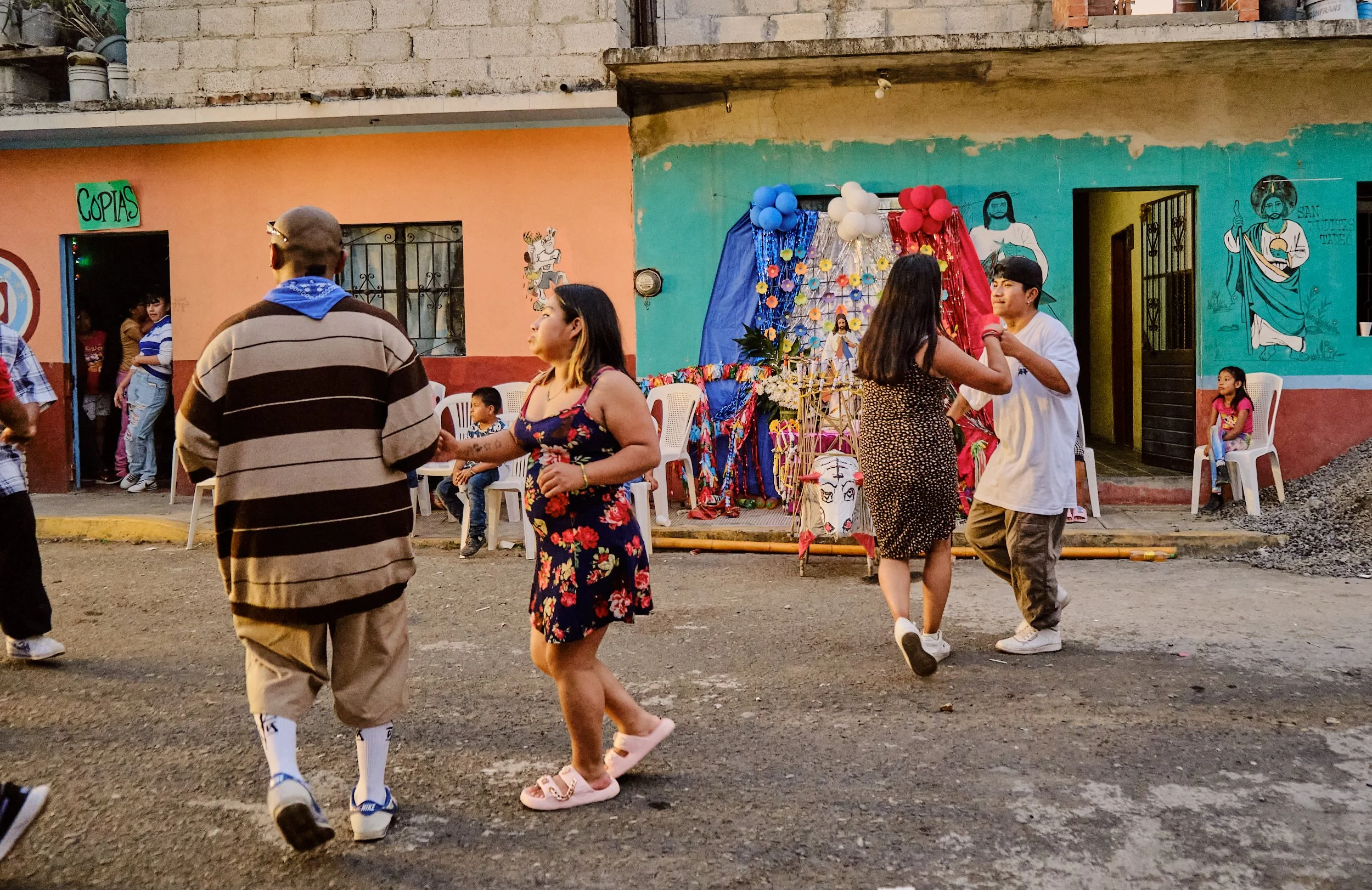 Personas bailando en una celebración en una calle, con decoraciones de flores, globos y una imagen religiosa en el fondo, en un vecindario de colores vivos.