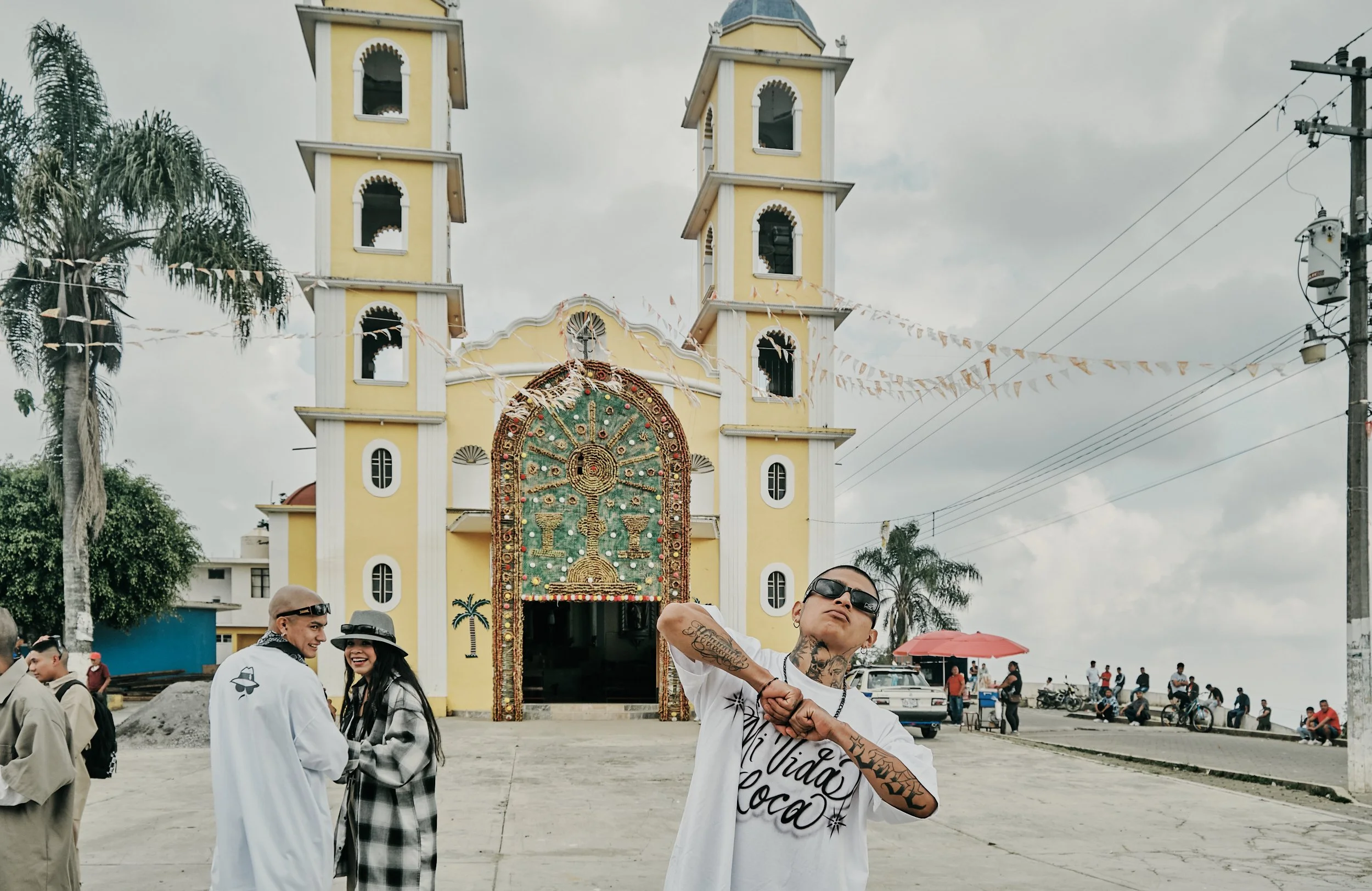 Grupo de personas en la plaza frente a una iglesia color amarilla con dos torres. Algunas personas están sonriendo y una está haciendo un gesto con su mano. El ambiente parece festivo con banderines decorativos y un cielo nublado.
