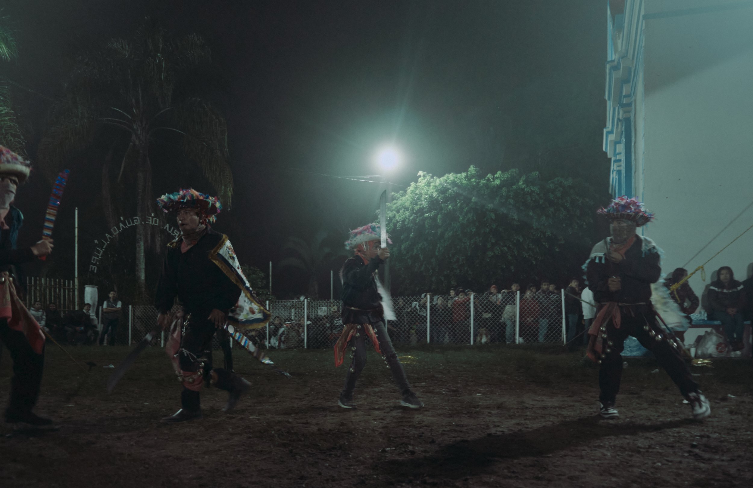 Personas disfrazadas bailando en una celebración nocturna con luna llena y árboles en el fondo.
