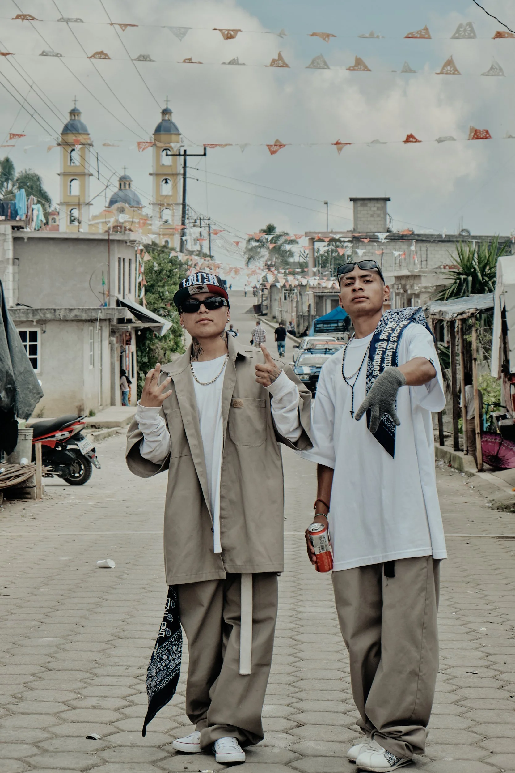 Dos jóvenes con estilo urbano posando en una calle de un barrio popular, con decoraciones de papel en el cielo, mientras algunos carros y personas se ven en el fondo.