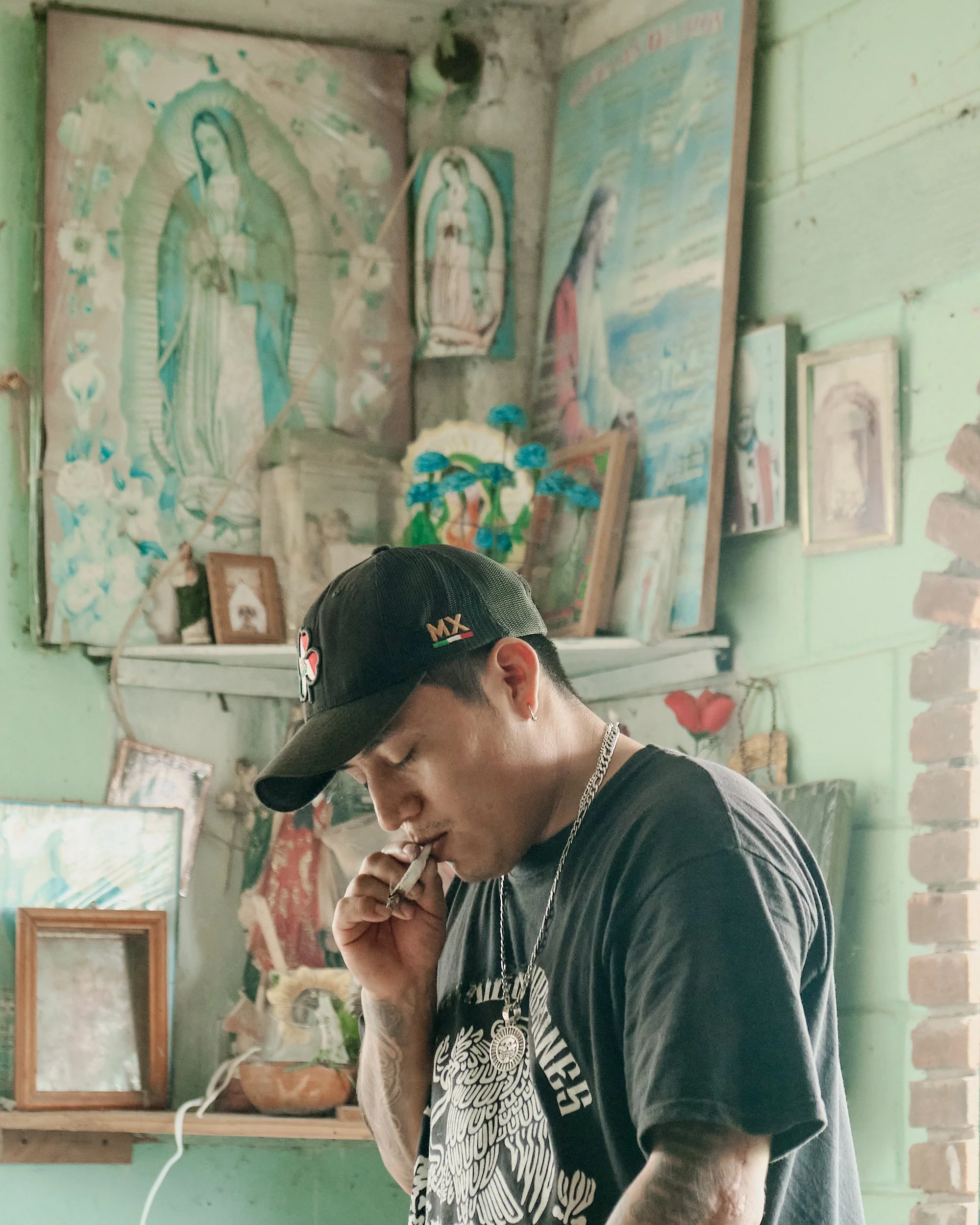 Joven con gorra negra y camiseta con diseño de ave, en un entorno con paredes verdes y decoraciones religiosas, sosteniendo un cigarro.