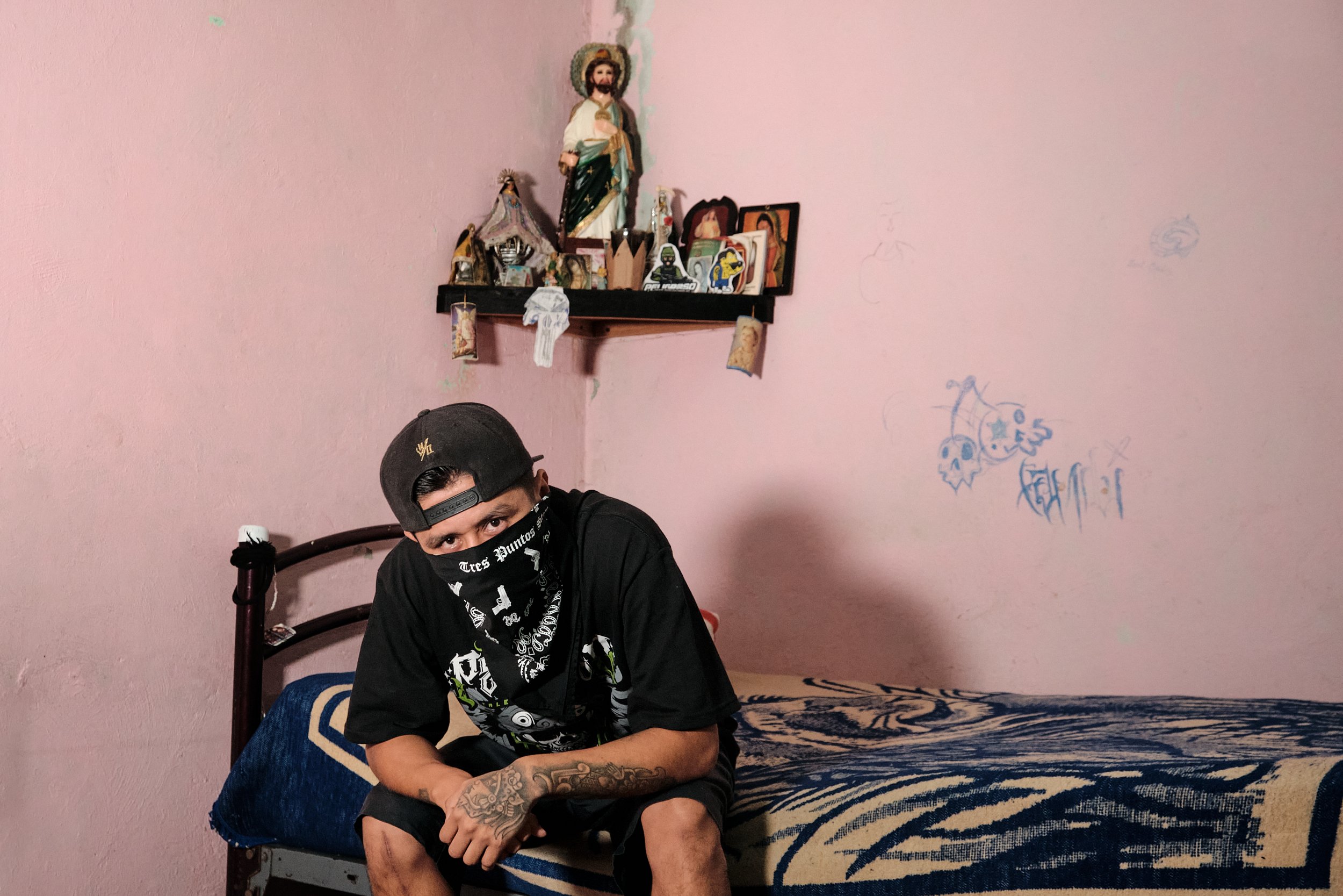 Joven con gorra negra, máscara negra con letras blancas, tatuajes en brazos, vestido con estampados en negro y verde, sentado en cama con colcha azul y crema, en habitación con paredes rosas y grafitis, y altar en la pared con figura de Jesús, fotogr
