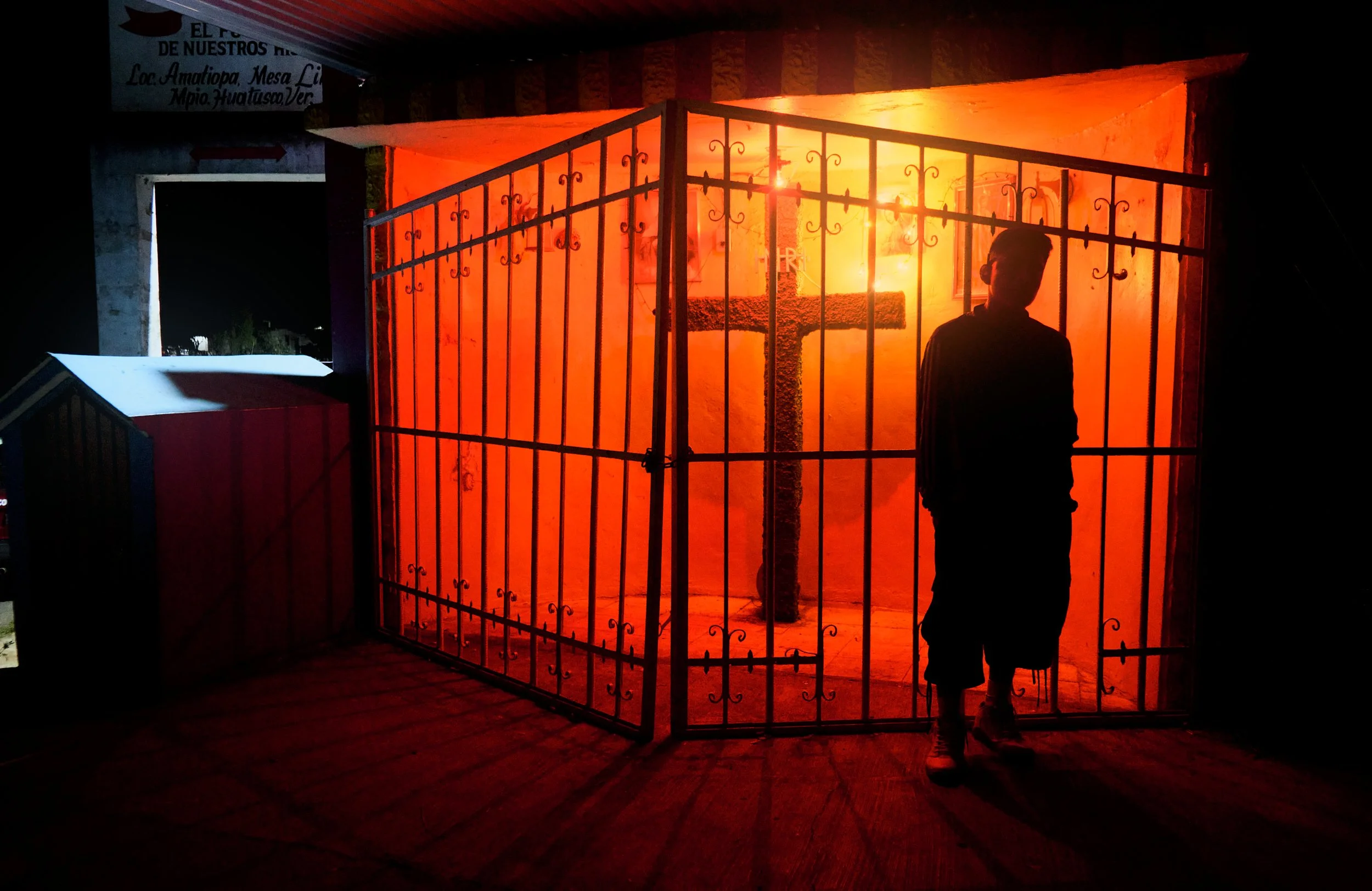 Silueta de una persona de pie frente a una puerta con reja de metal, iluminada con fuego o luz intensa de color naranja y rojo.