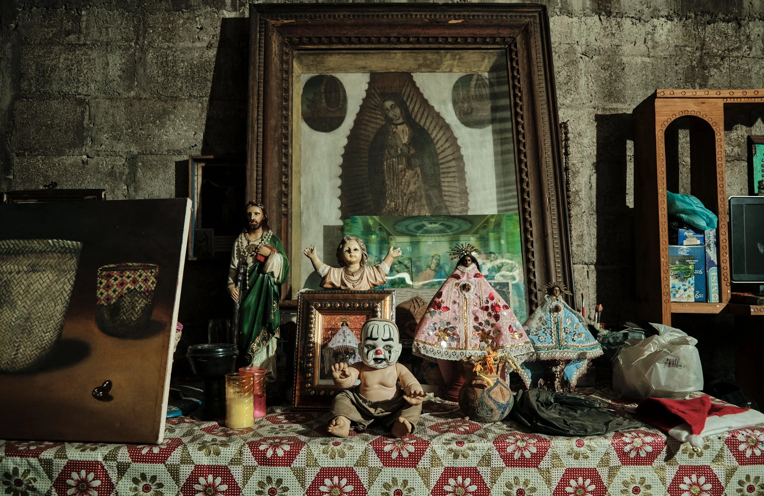 Muebles y objetos religiosos, incluyendo figurines de santos, una máscara de payaso y adornos textiles en una mesa con fondo de paredes de piedra