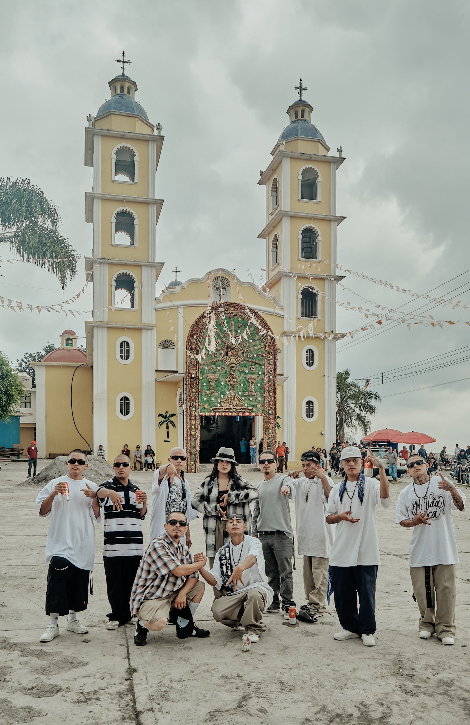 Un grupo de personas posando frente a una iglesia de papel maché decorada con símbolos religiosos, con nubes grises en el cielo, en un evento religioso en una plaza al aire libre.