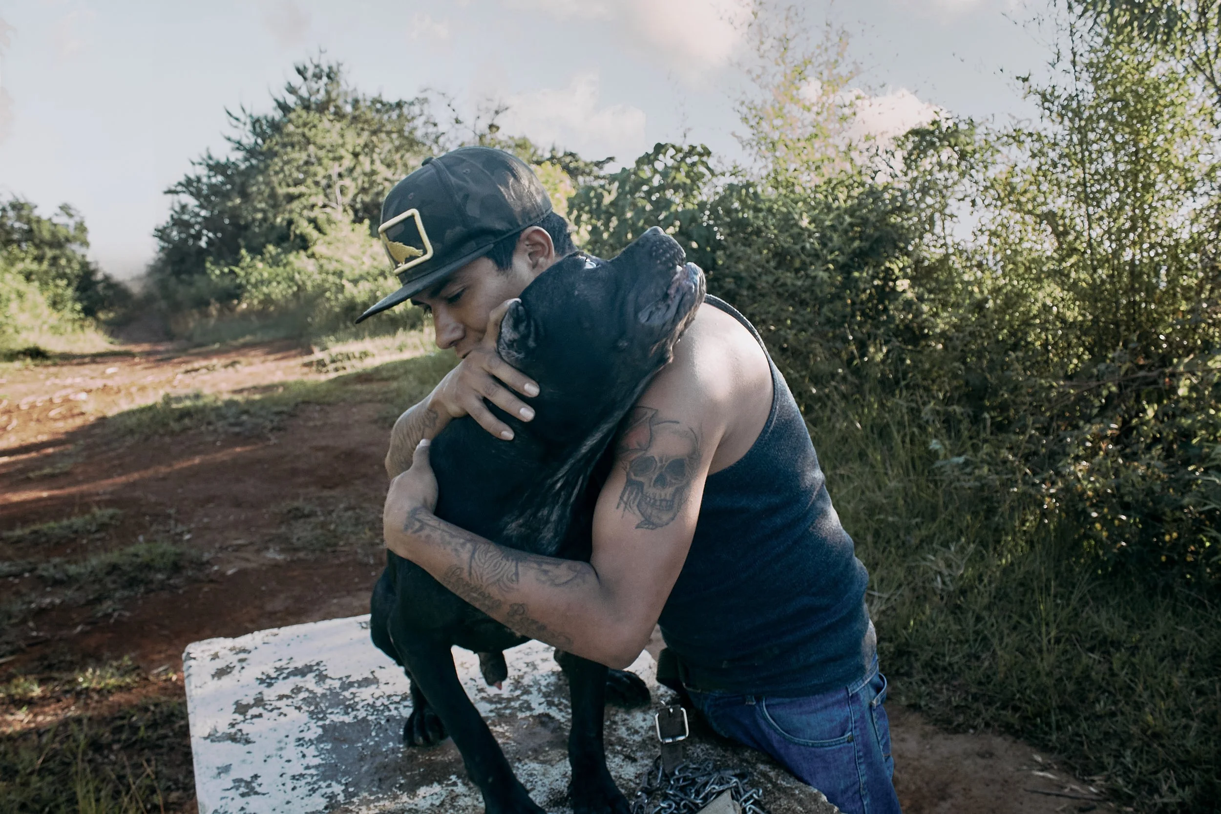 Un hombre con tatuajes abrazando a un perro negro en un entorno al aire libre con árboles y césped.