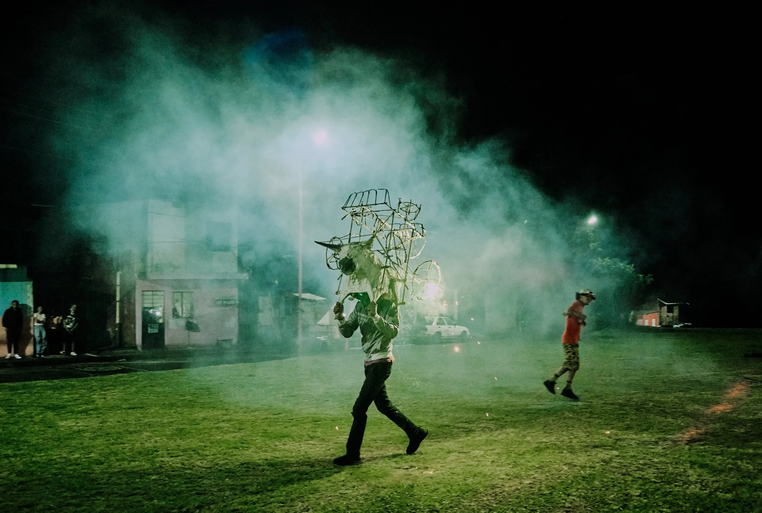 Persona sosteniendo una estructura de alambre y tela en un campo con humo y polvo, varias personas observando en el fondo, noche con iluminación artificial.