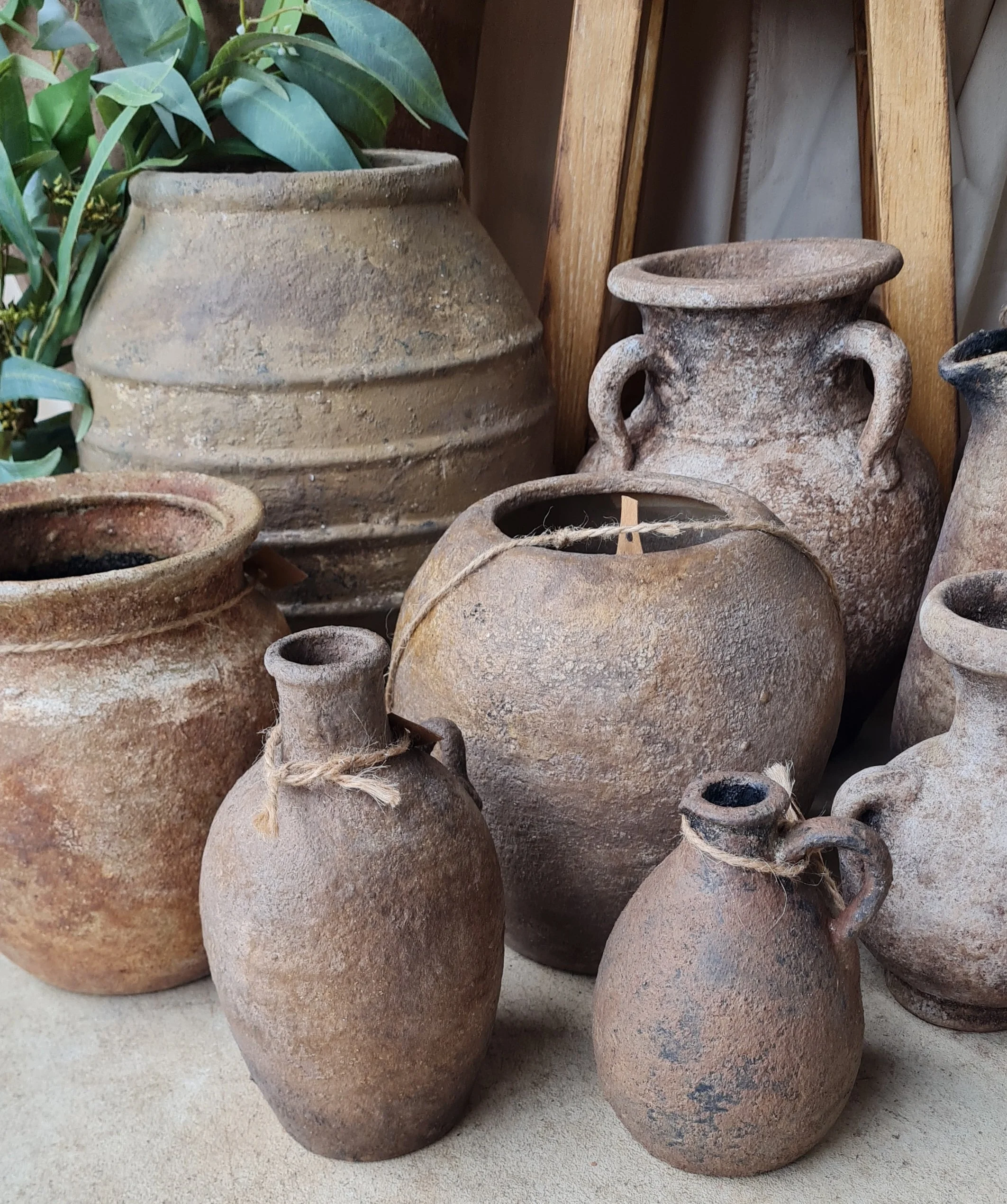 Collection of old, weathered clay pots and vases in earthy tones, some with jute rope handles, arranged outdoors on a surface with a wooden background.