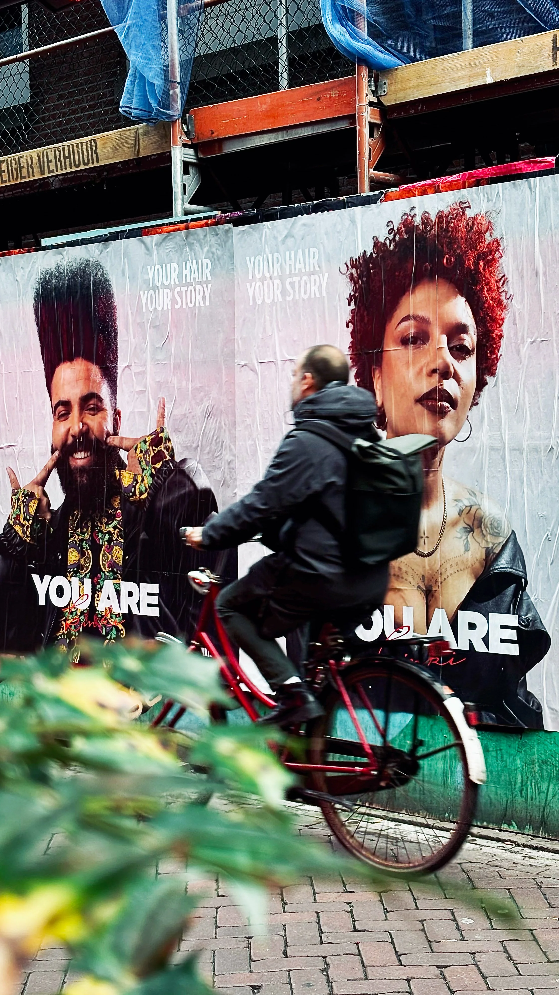 A person riding a red bicycle passes a large advertisement poster featuring two diverse models with vibrant hair styles. The poster has the text 'YOUR HAIR YOUR STORY' and 'YOU ARE' prominently displayed.