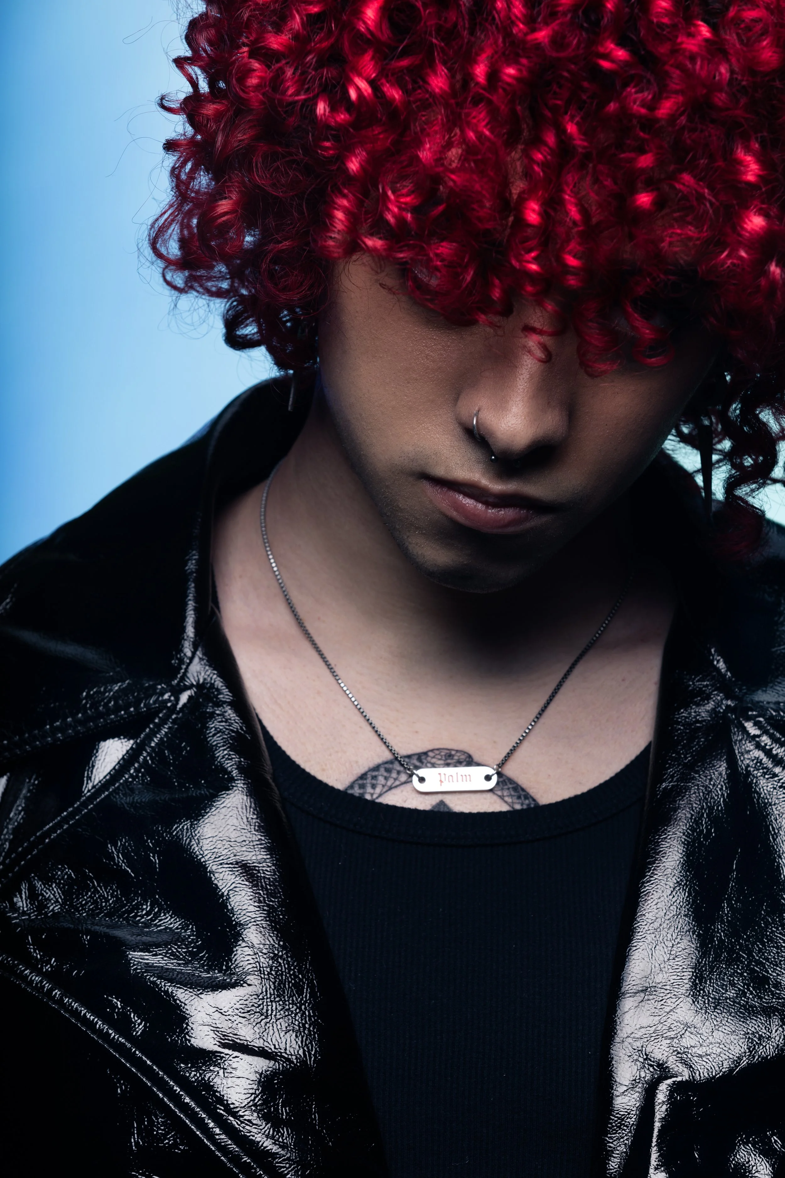 A person with vibrant curly red hair covering part of their face, wearing a black leather jacket, a black shirt, a silver necklace, and a septum ring, with a blue background.