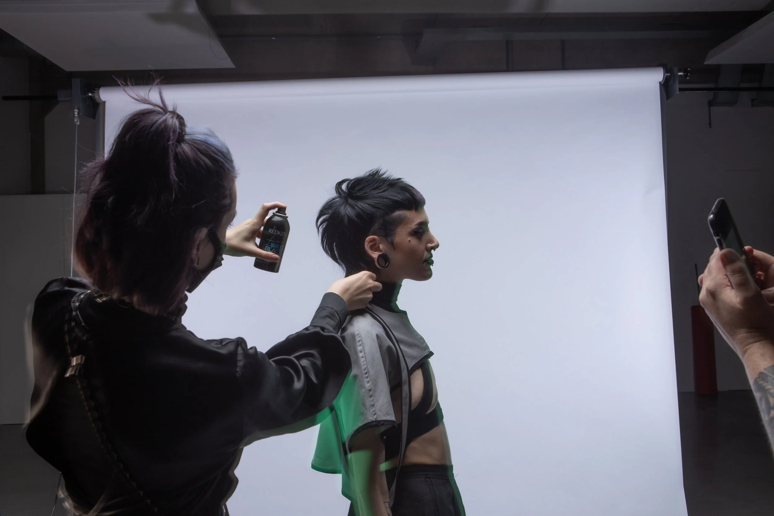 A fashion model with short black hair and large earrings poses against a white backdrop while a stylist applies hair spray and a person on the right captures the scene with a smartphone.