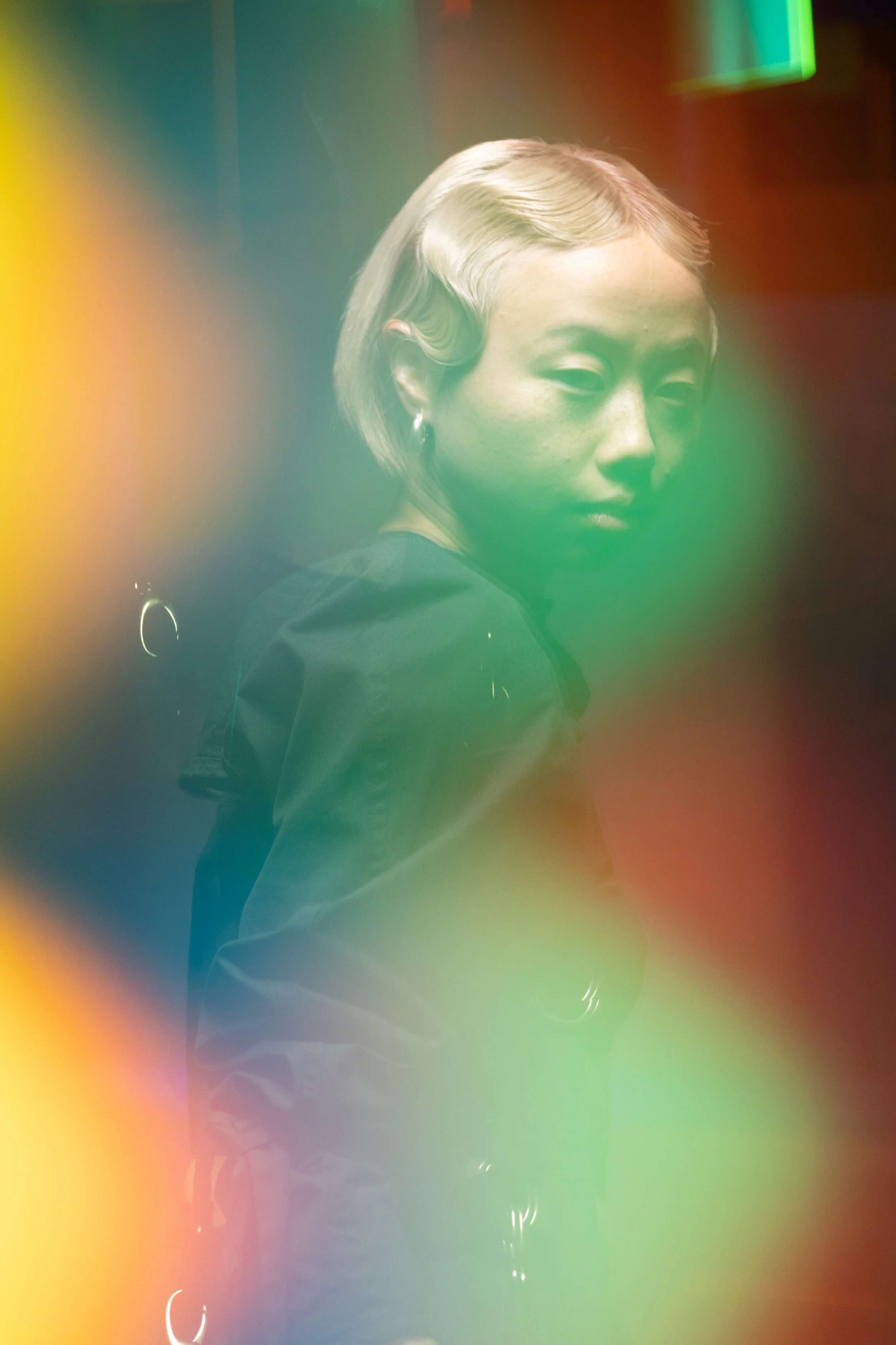 A woman with short blonde hair, wearing earrings, is seen through colorful, blurred light overlays.