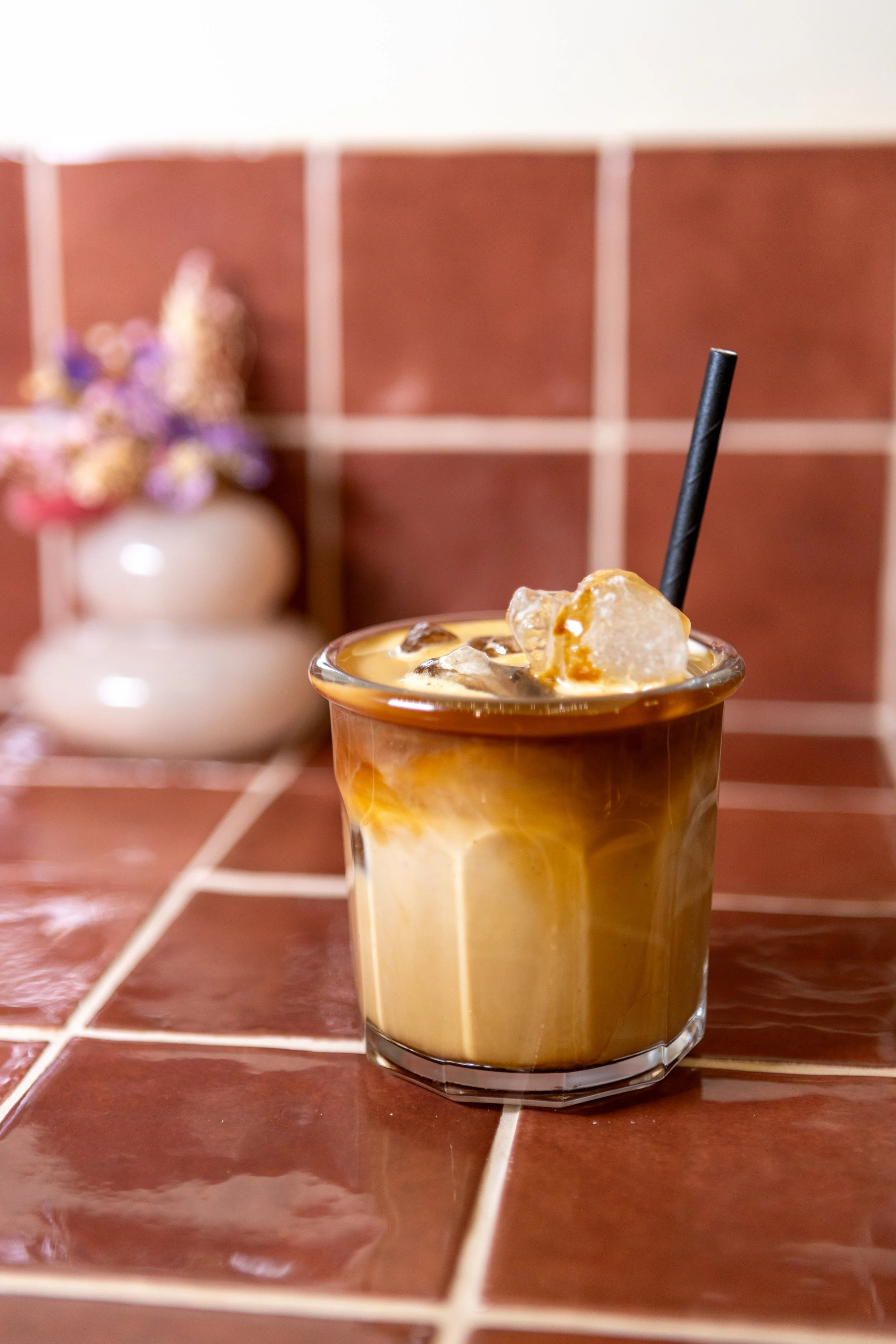 A glass of iced coffee with milk and caramel swirls, on a tiled surface with a blurred flower vase in the background.