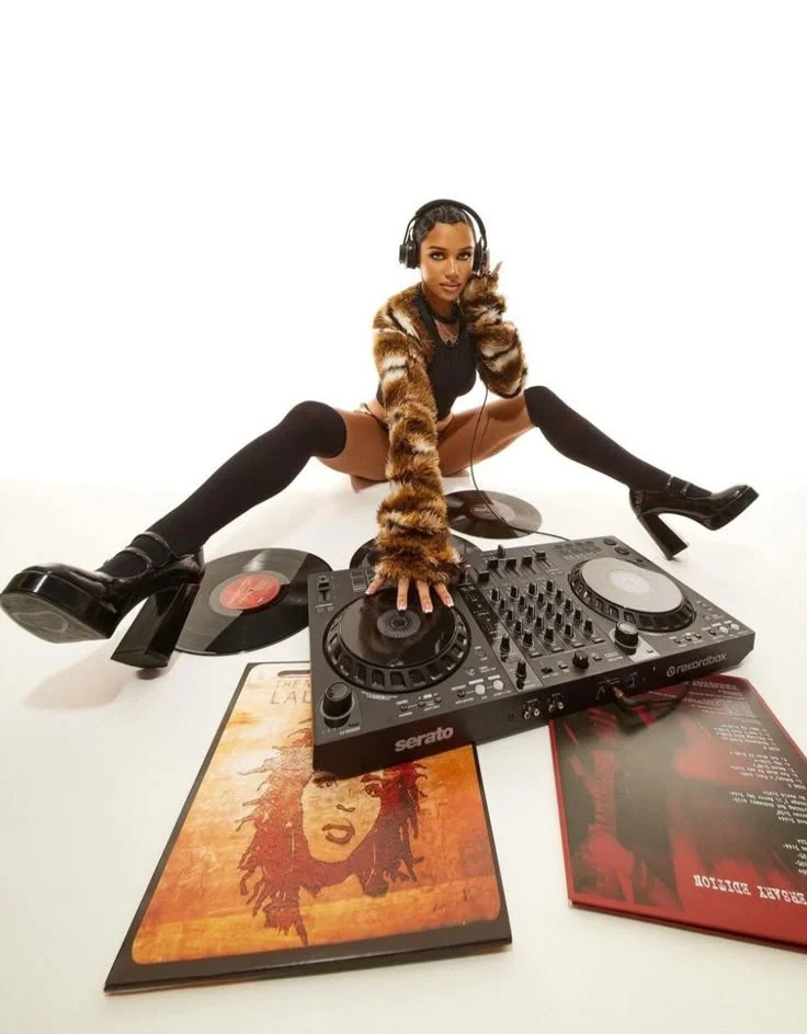A woman with dark hair, wearing a leopard print jacket, black thigh-high socks, and black high-heeled boots, sitting on the floor with legs apart, using a DJ controller surrounded by vinyl records and album covers on a plain white background.