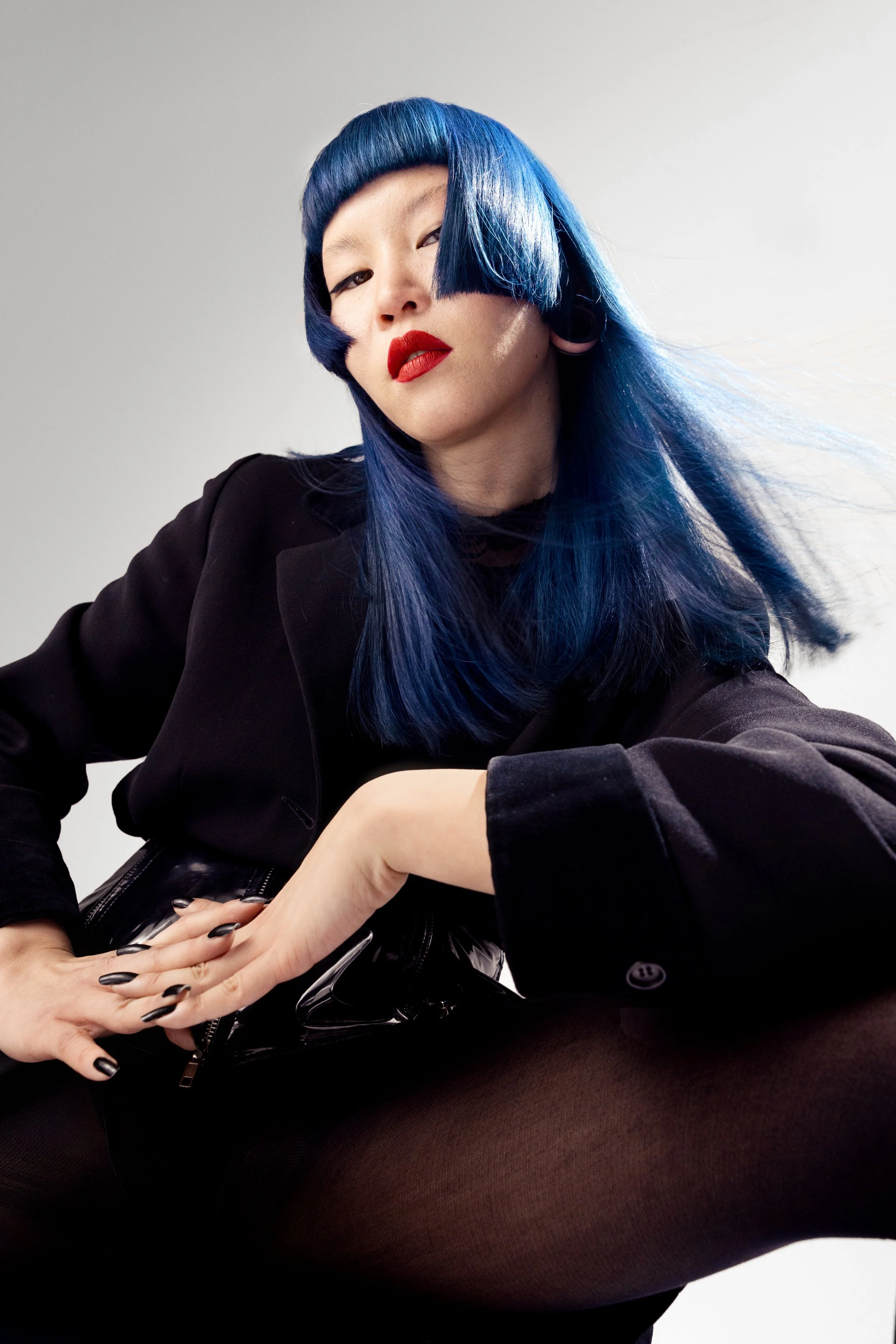 A woman with blue hair and red lipstick poses confidently in black clothing against a plain background.