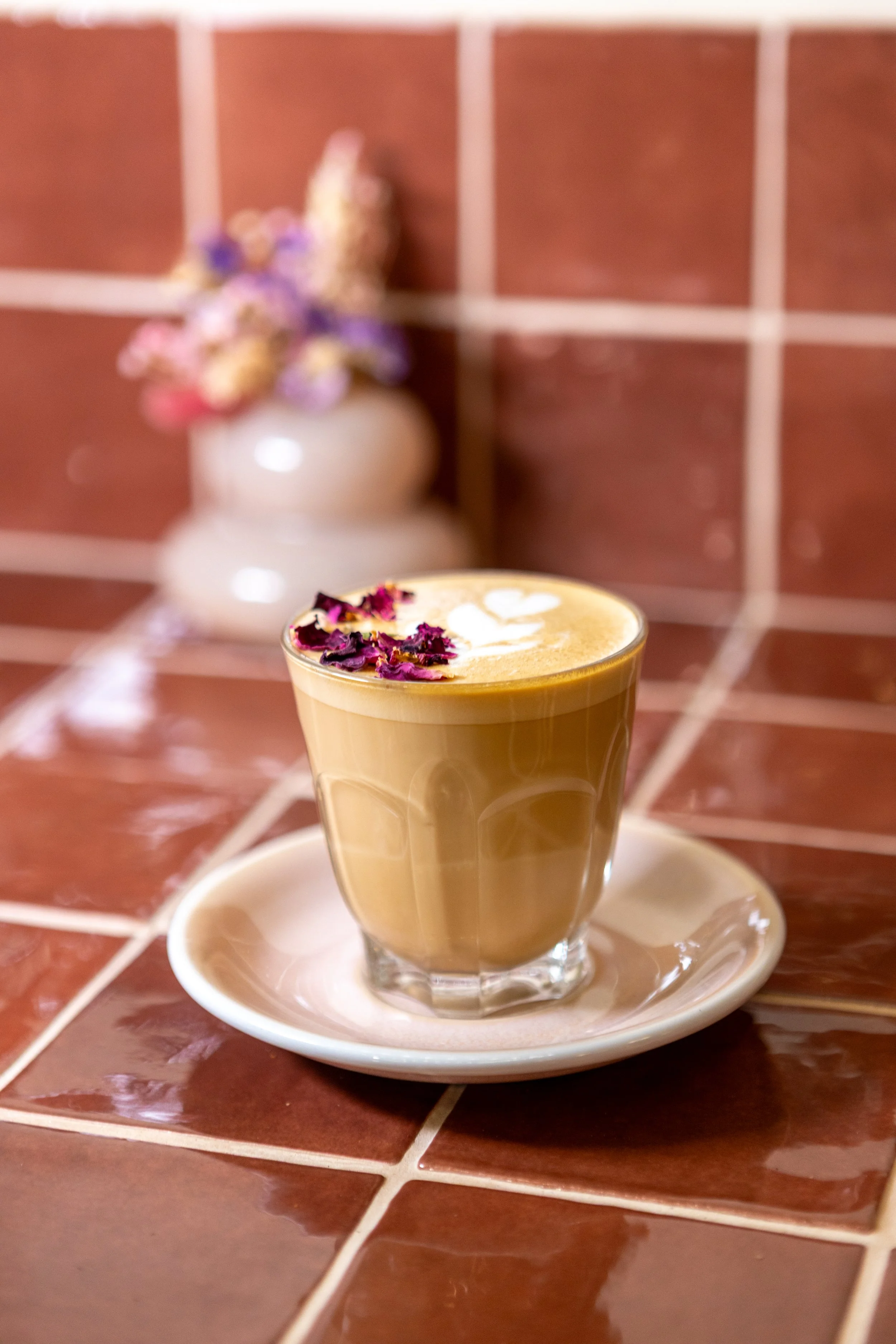 A latte with foam art and dried flower petals on top, in a clear glass cup on a white saucer, on a brick-tiled surface with a blurred decorative vase and flowers in the background.