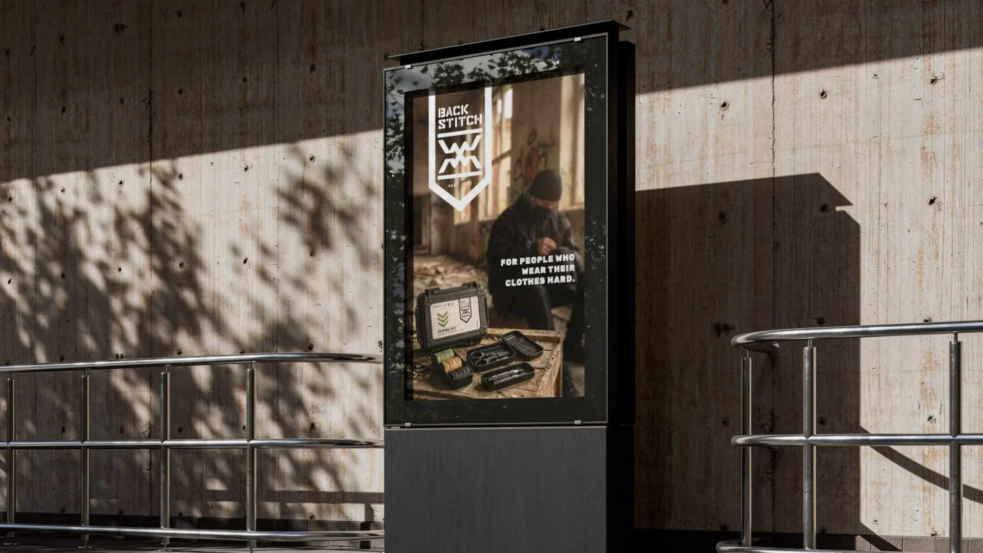 A digital advertisement for a clothing brand called Back Stitch, featuring a man sitting in an abandoned building with graffiti, wearing a beanie, and surrounded by sewing and crafting tools on a wooden table. The ad text reads: "For people who wear 