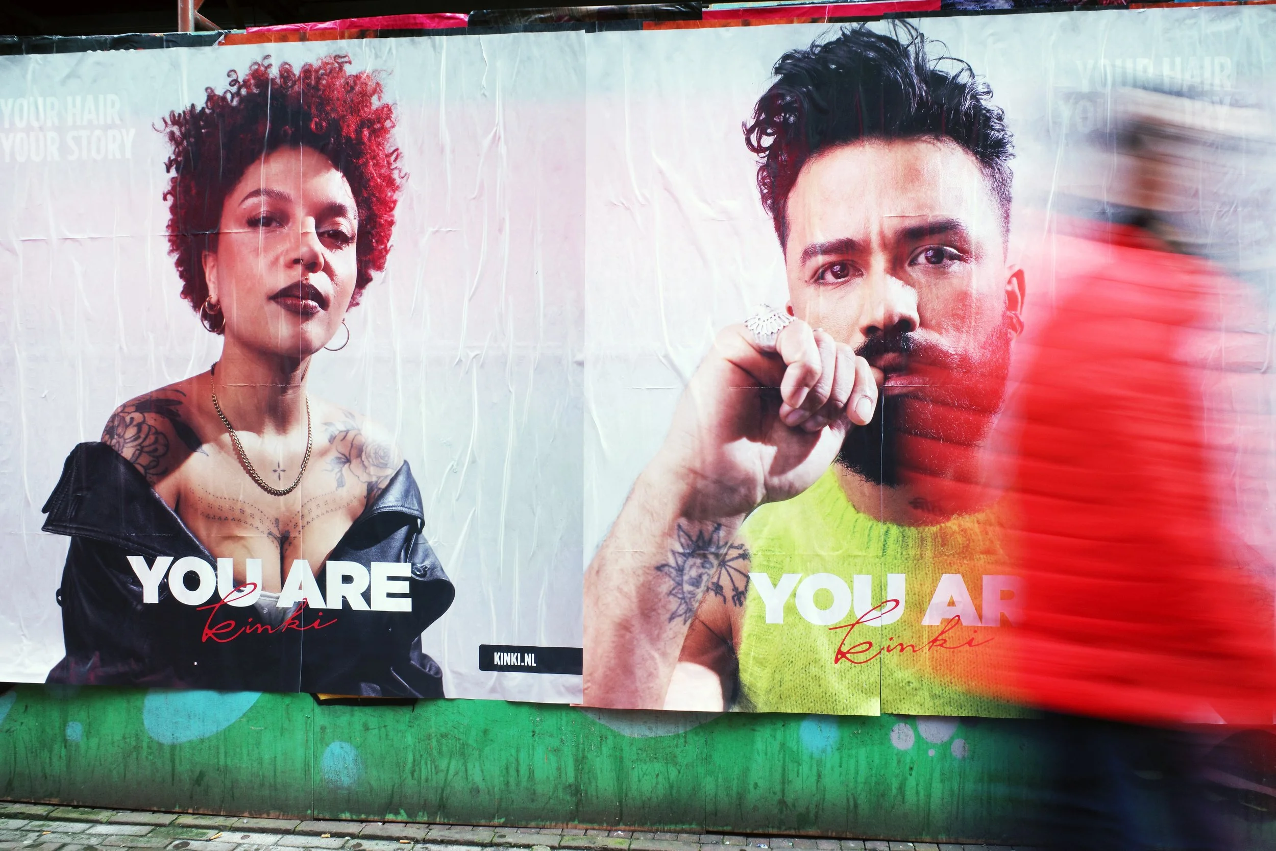 A large billboard featuring a woman with curly red hair and tattoos on her shoulders, wearing a black leather jacket, and a man with dark hair and a beard, wearing a bright green shirt, with the text 'You Are' and 'Reinki' on the billboard.