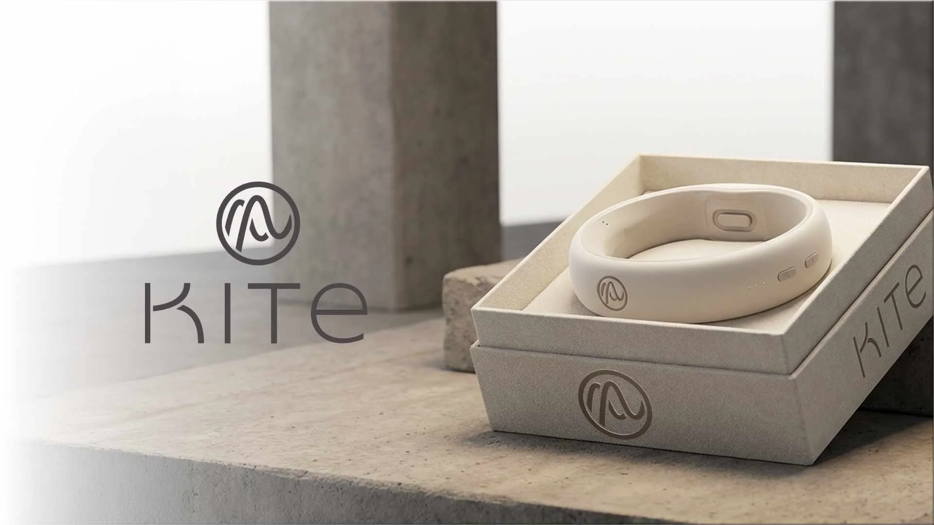 A white wearable device, possibly a fitness or health tracker, is placed inside an open box with the brand name 'KITE' and logo visible on both the device and the box. The scene features a minimalist background with neutral-colored stones and a woode