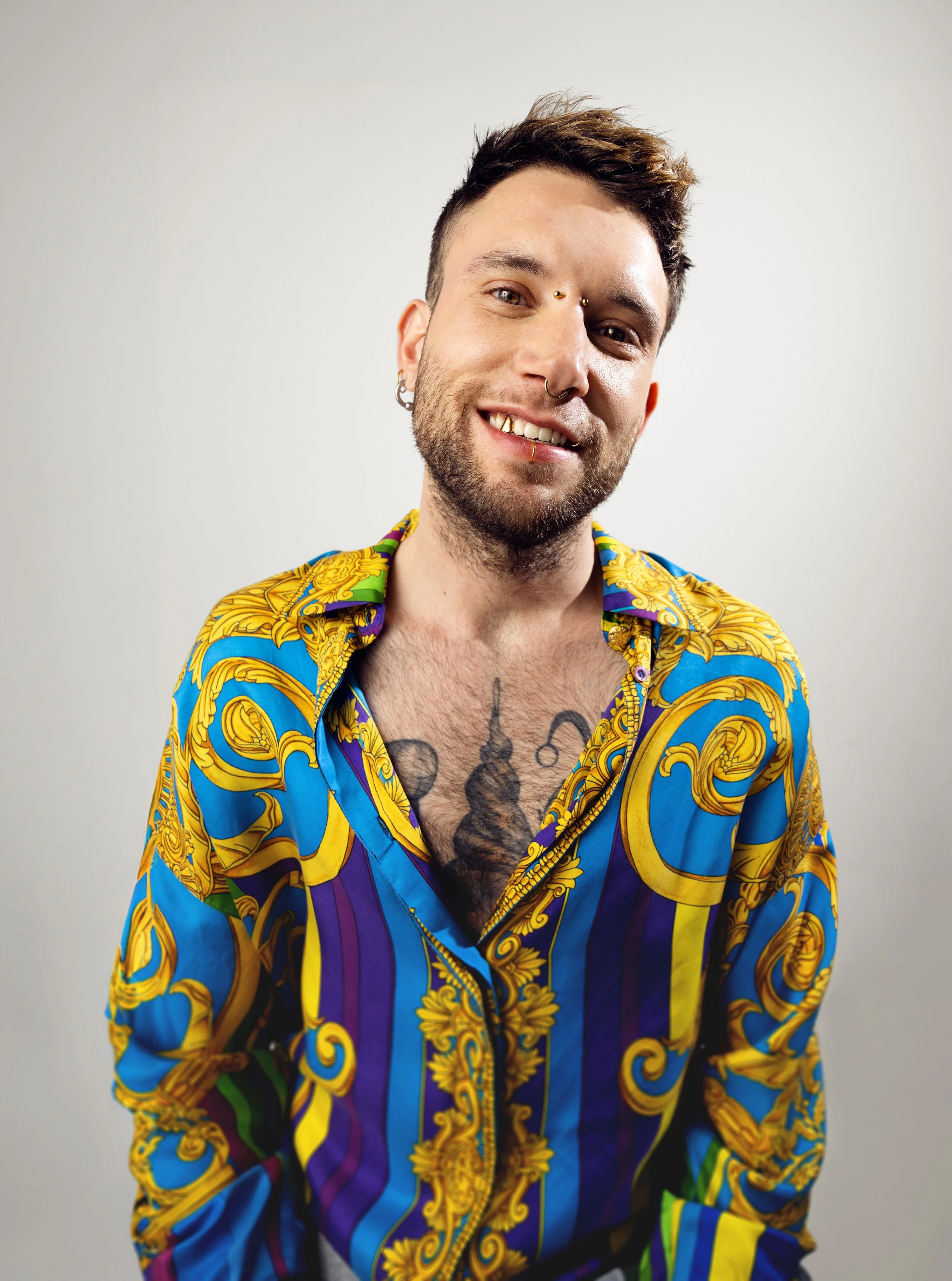 A smiling man with tattoos, piercings, and short styled hair wearing a colorful, patterned shirt against a plain background.