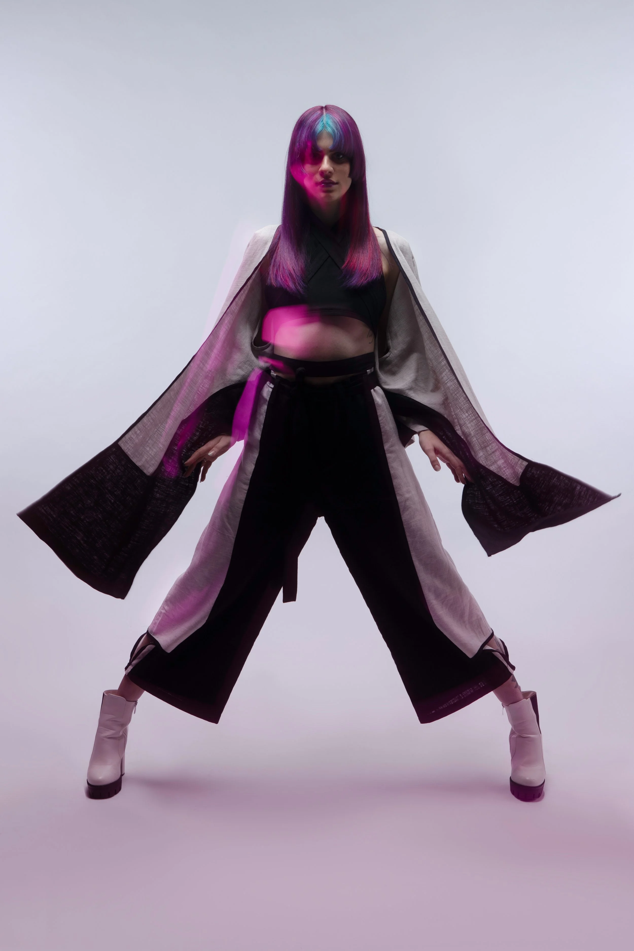 Fashion model standing confidently with legs apart, wearing a long, open coat over a black crop top and wide-leg pants, with colorful hair and white ankle boots, against a plain white background.