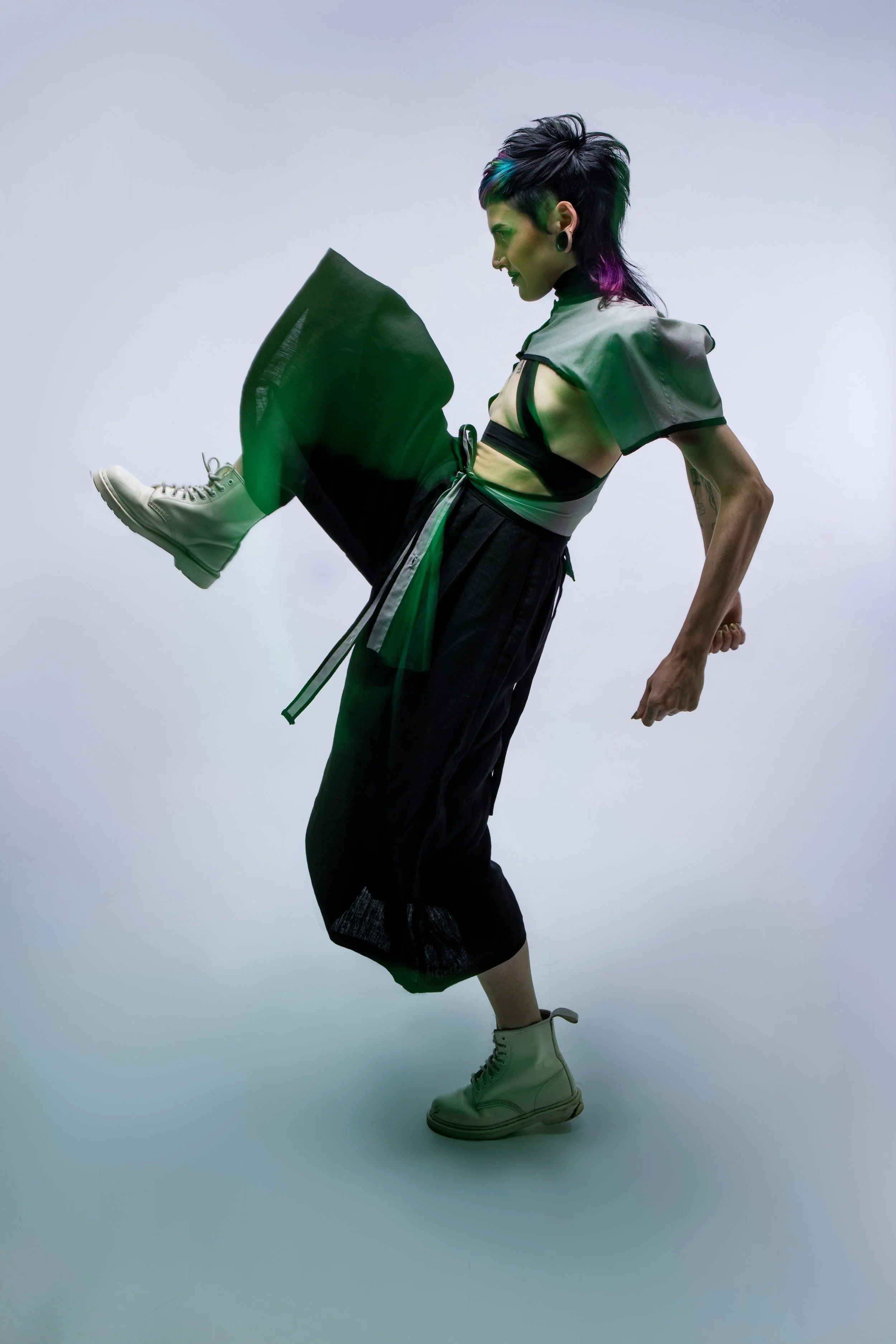 Person with short, spiky hair and multiple piercings in a dynamic pose, wearing a layered outfit with a cropped top, wide-leg pants, and high-top shoes, against a plain background.
