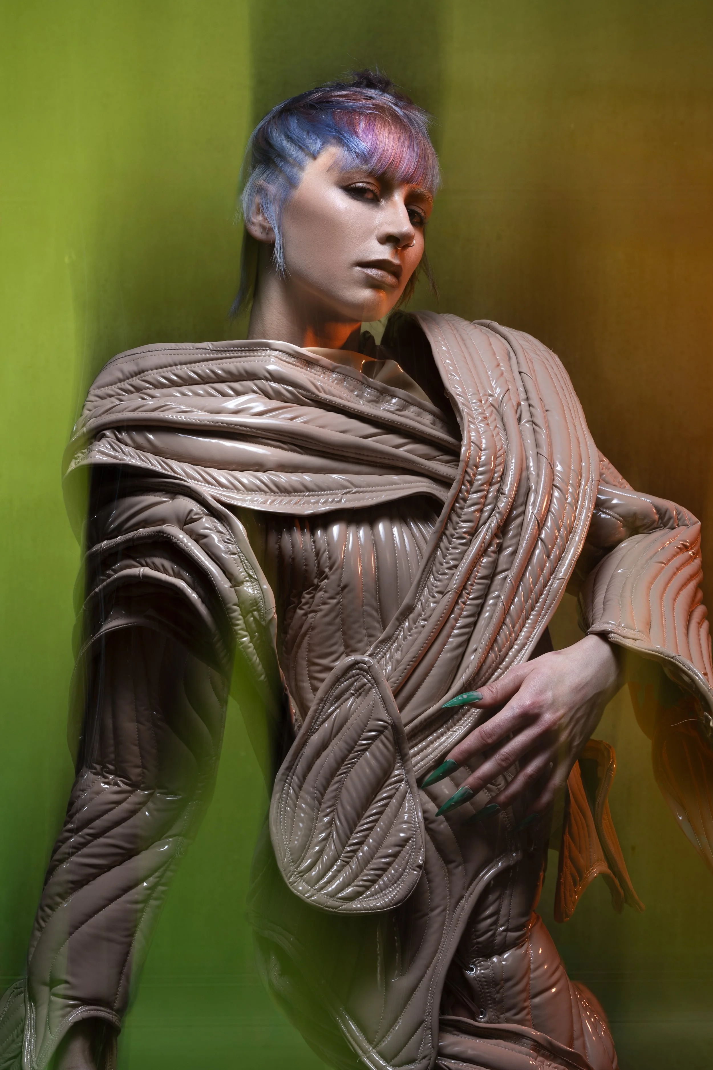 Person with short, multi-colored hair wearing a shiny, beige, quilted jacket, standing against a green background.