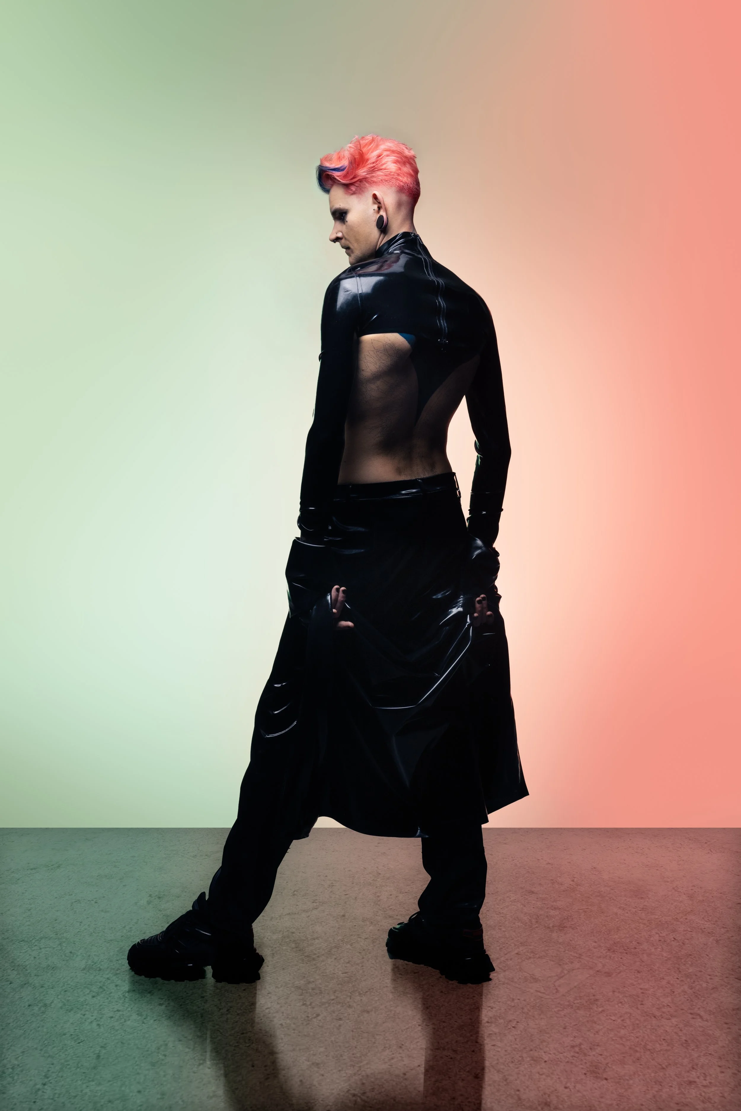 A person with pink hair and wearing black clothing, standing against a background with a gradient from green to pink.