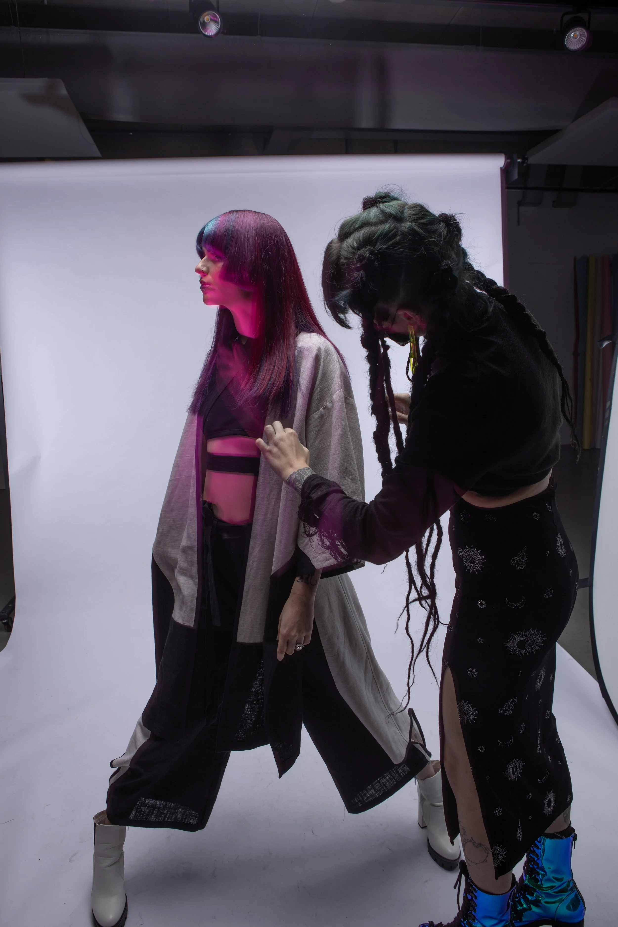A woman in a gray overcoat and black pants standing against a white backdrop, with another person adjusting her coat. The woman has straight, dark hair with colorful bangs, and is dressed in fashion-forward clothing, likely in a photoshoot studio setting.