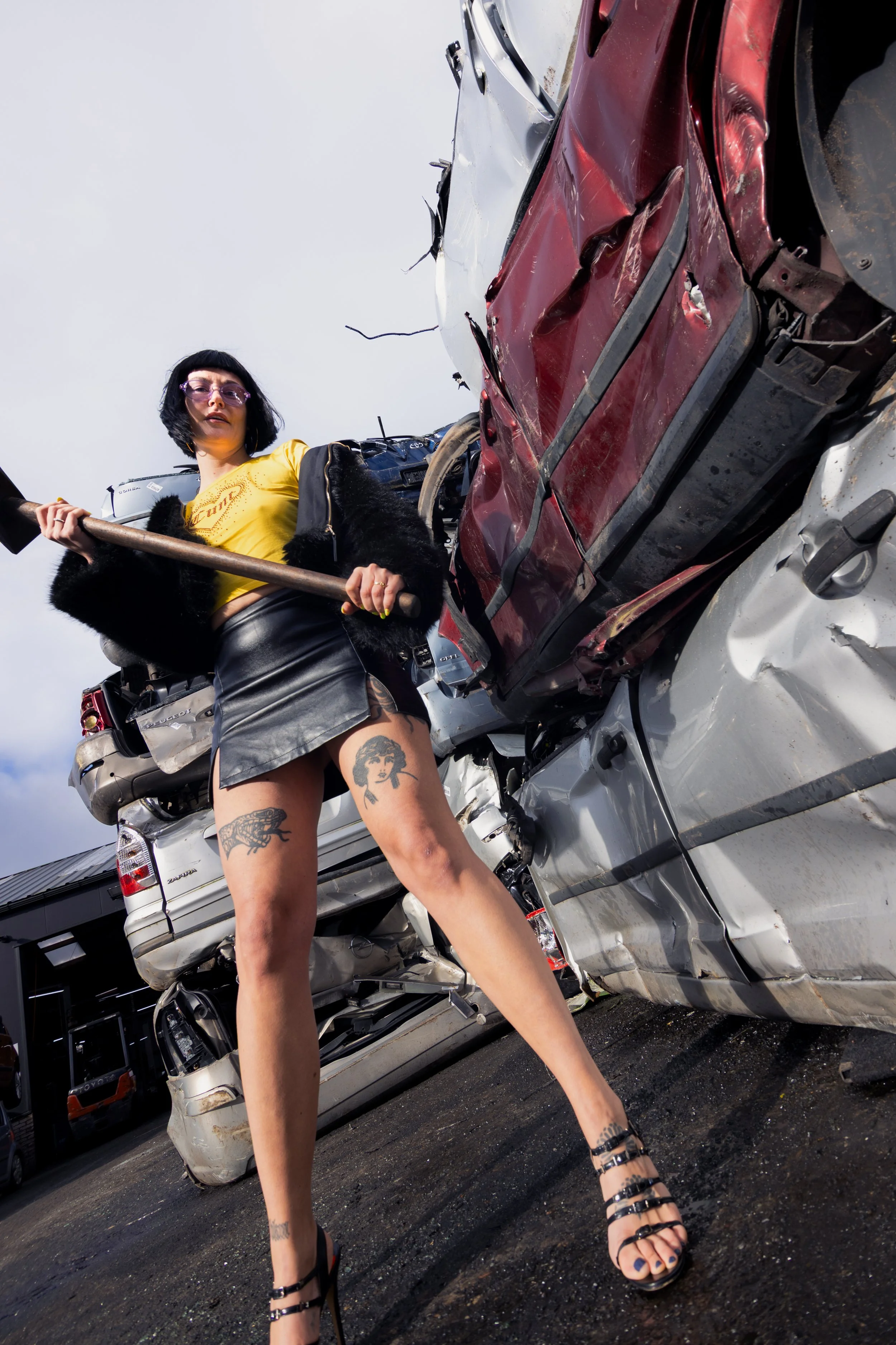 Woman with tattoos on her legs standing amidst a car crash, holding a sledgehammer, with damaged cars in the background.