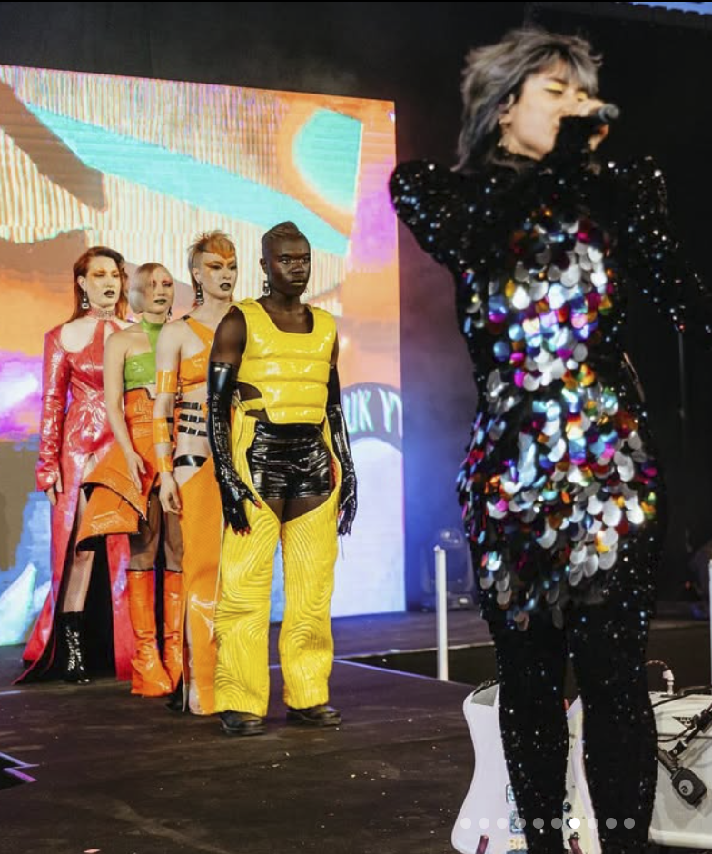A performer in a black sequin outfit decorated with large, shiny multicolored sequins is singing into a microphone; behind them, five models dressed in colorful, futuristic outfits are standing in a line on a runway, with a vibrant, abstract digital backdrop.