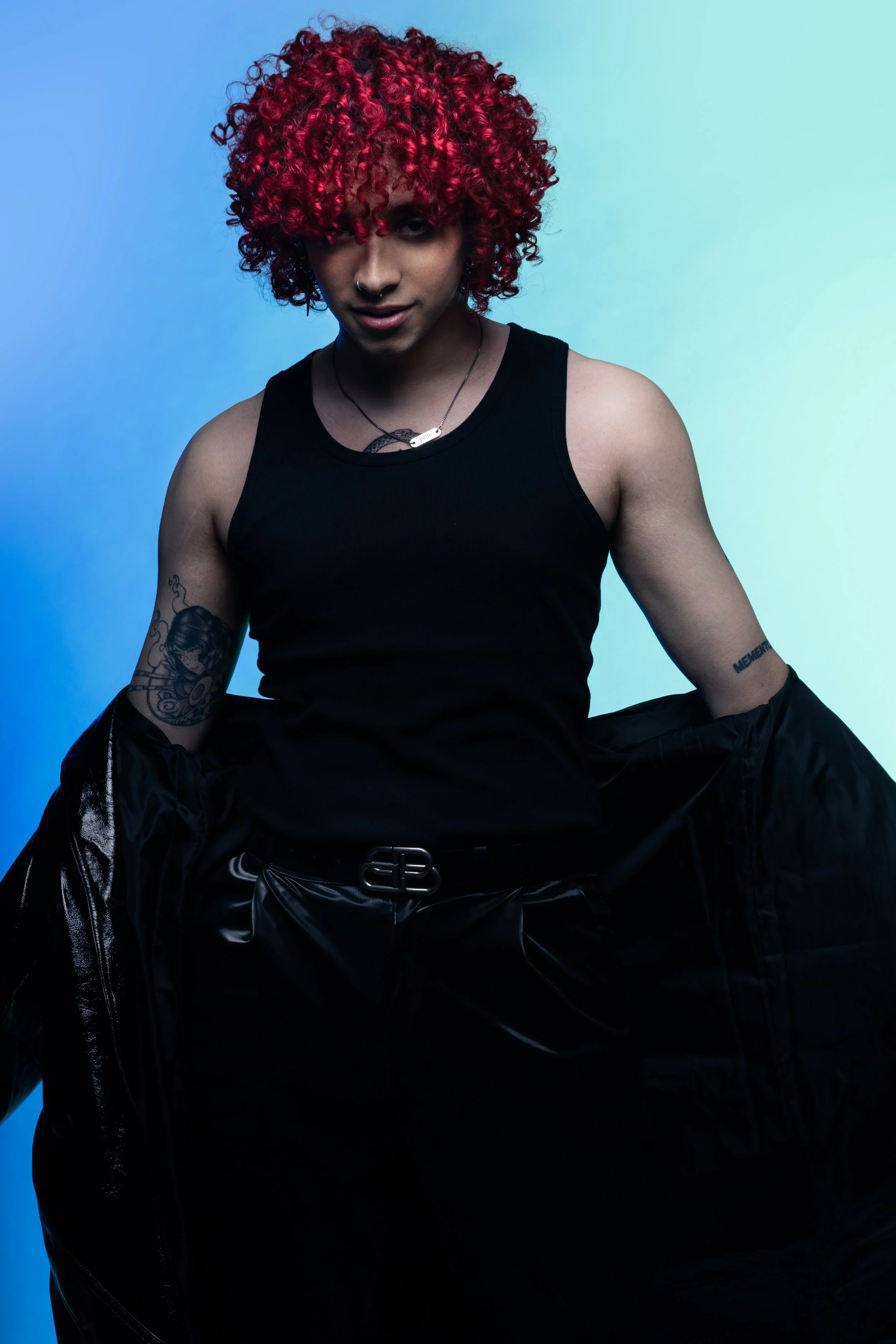 Person with curly bright red hair wearing a black sleeveless top, black pants, and tattoos, standing against a blue and teal gradient background.
