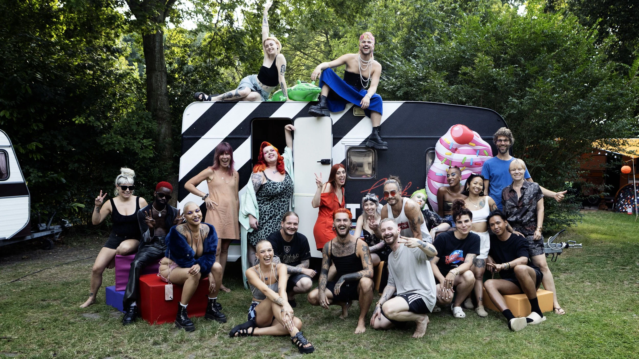 Group of diverse people celebrating outdoors with a camper trailer in a wooded area, some sitting on colorful blocks, others standing or sitting near the camper, with some on top of it, all smiling and making peace signs, dressed in casual and colorful clothing.