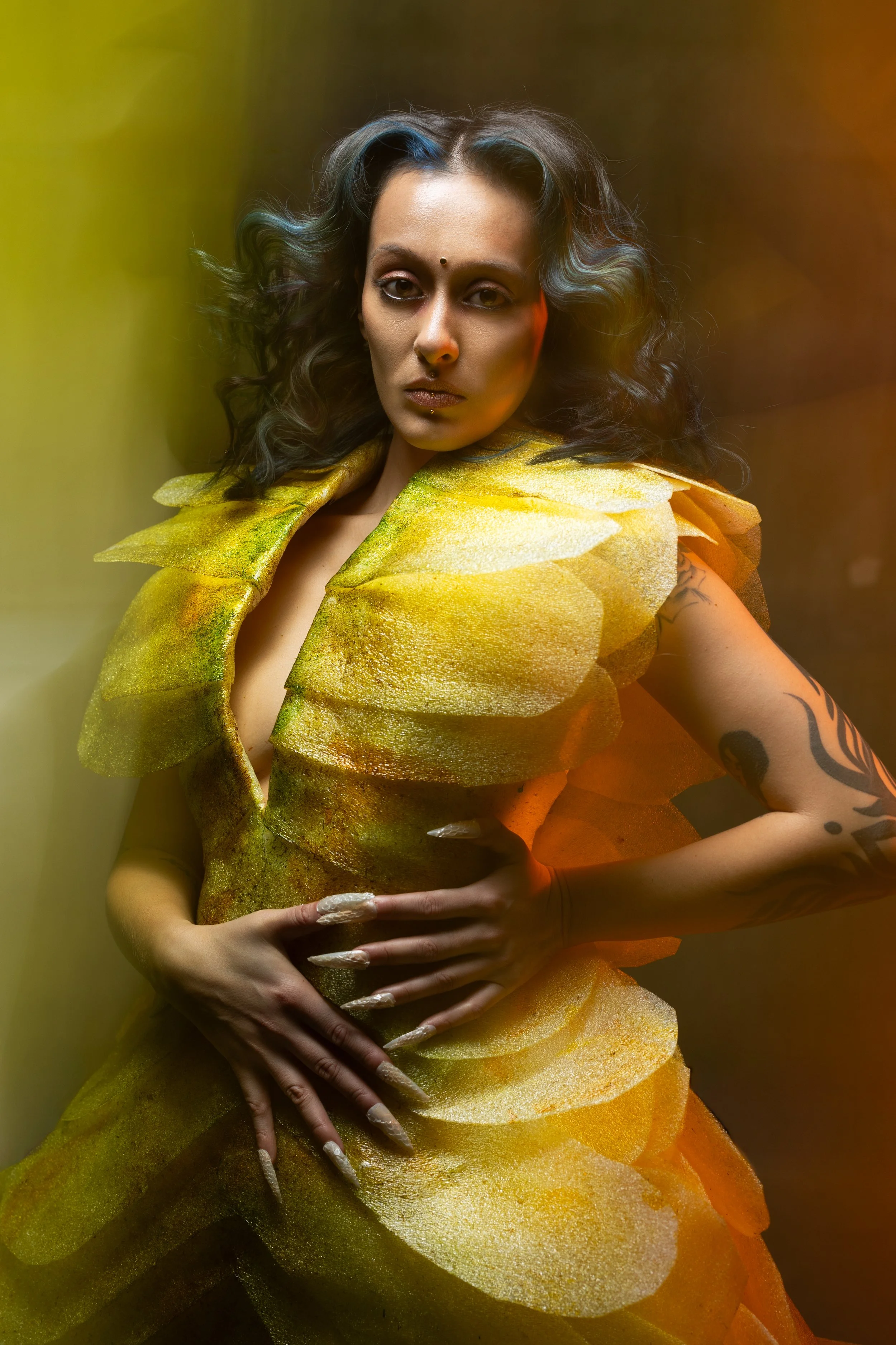 A woman with curly hair and tattoos wearing a dress made of layered, yellow-green petal-like fabric, standing with her hands on her stomach against a multicolored background.