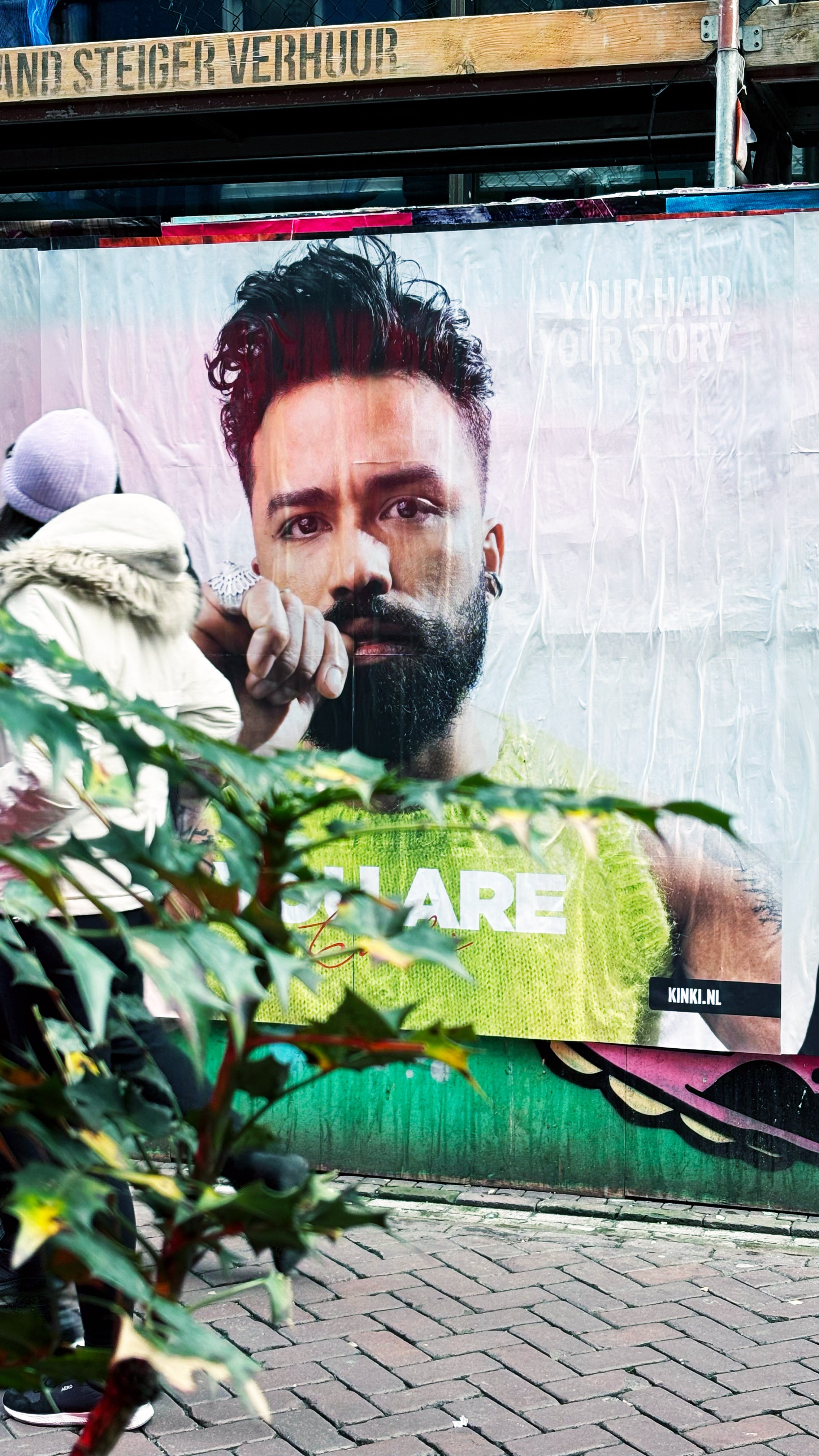 A large outdoor advertisement featuring a close-up of a bearded man with styled hair, wearing a bright green shirt. Behind the poster, part of a building with wooden siding and a metal railing is visible. The ground is paved with bricks, and some gre