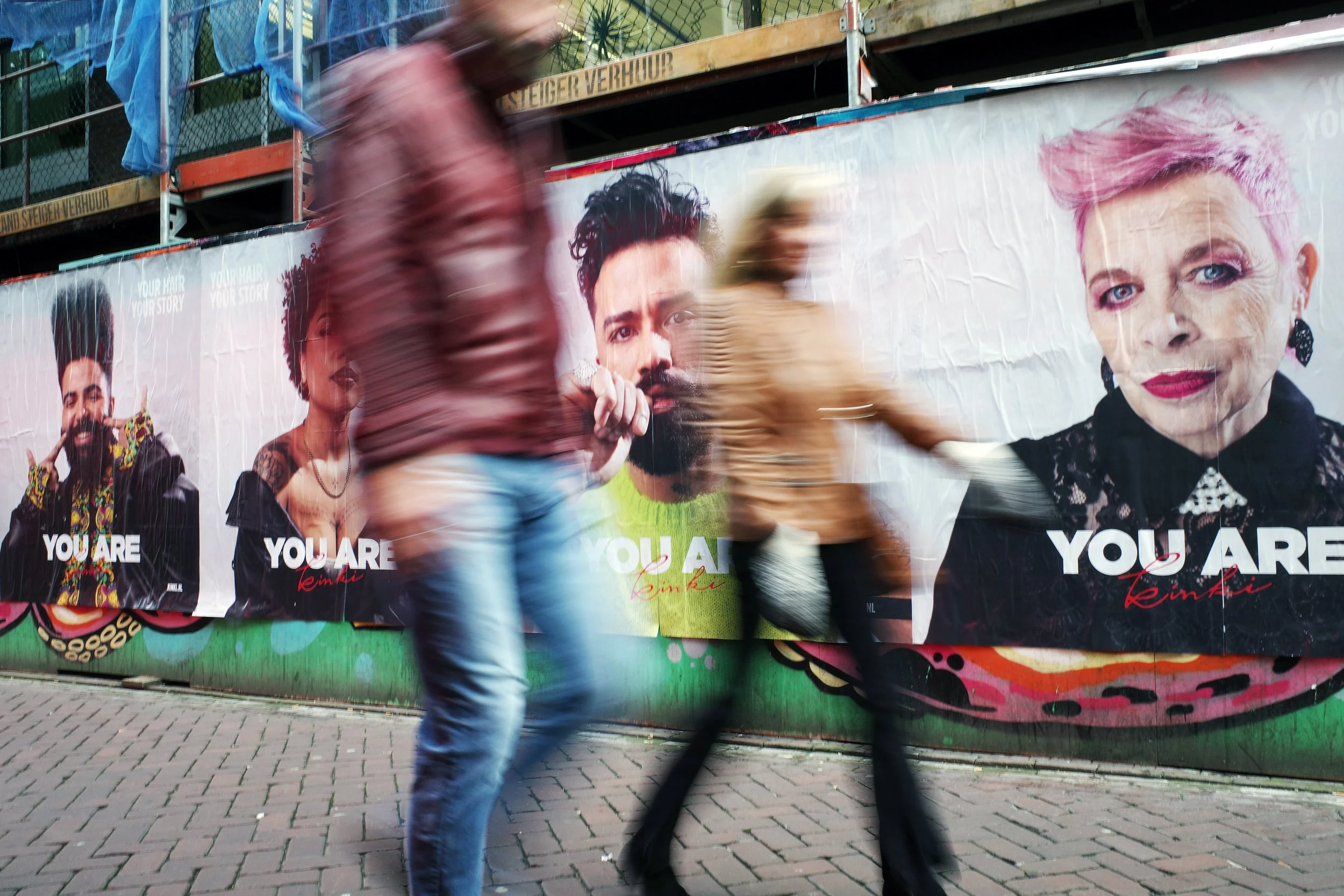 People walking past colorful posters on a city street, some motion blurred