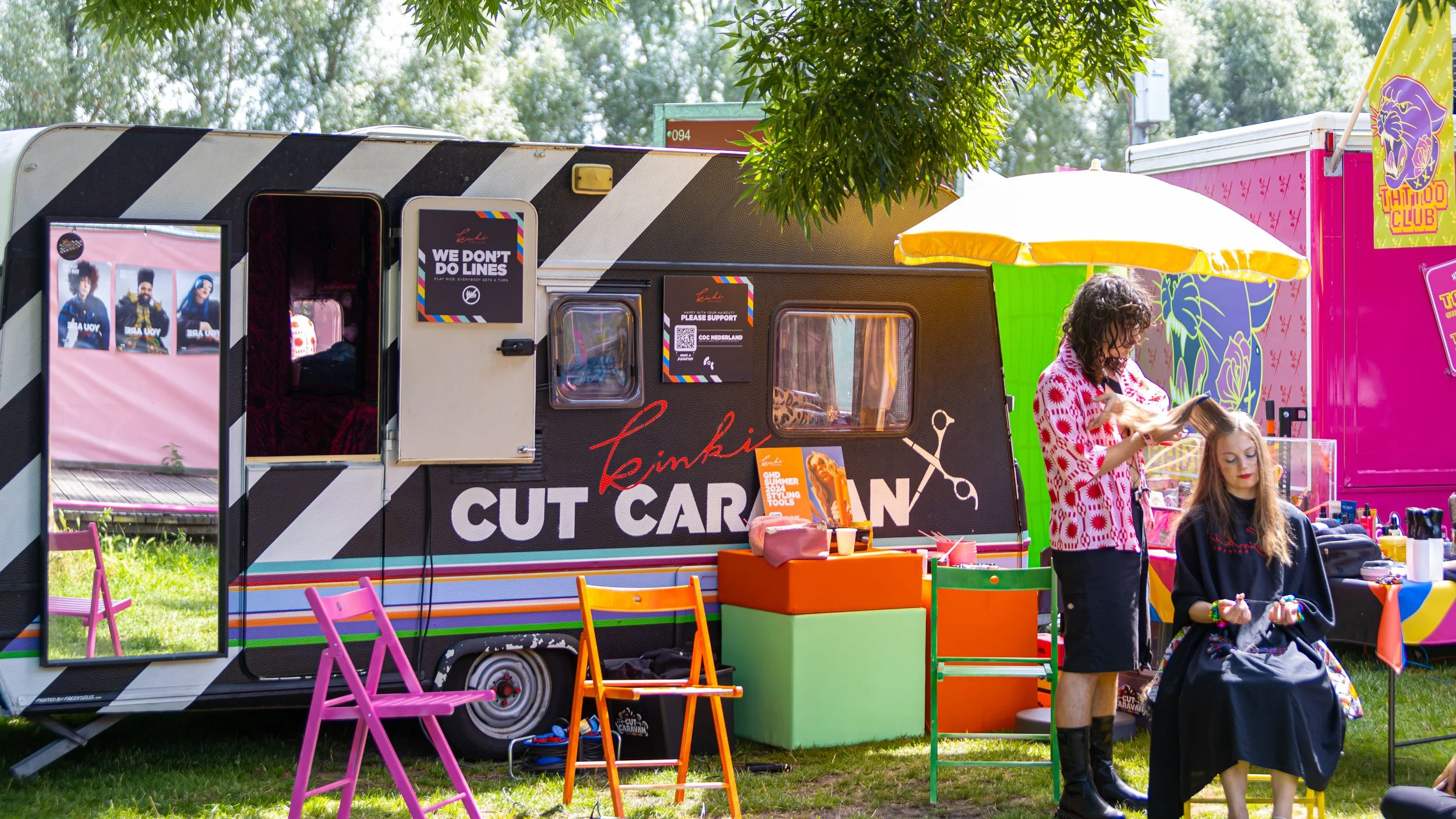 A woman getting her hair styled at a colorful outdoor hair salon beside a black trailer with diagonal stripes, pink chairs, and a yellow umbrella, with another woman sitting.