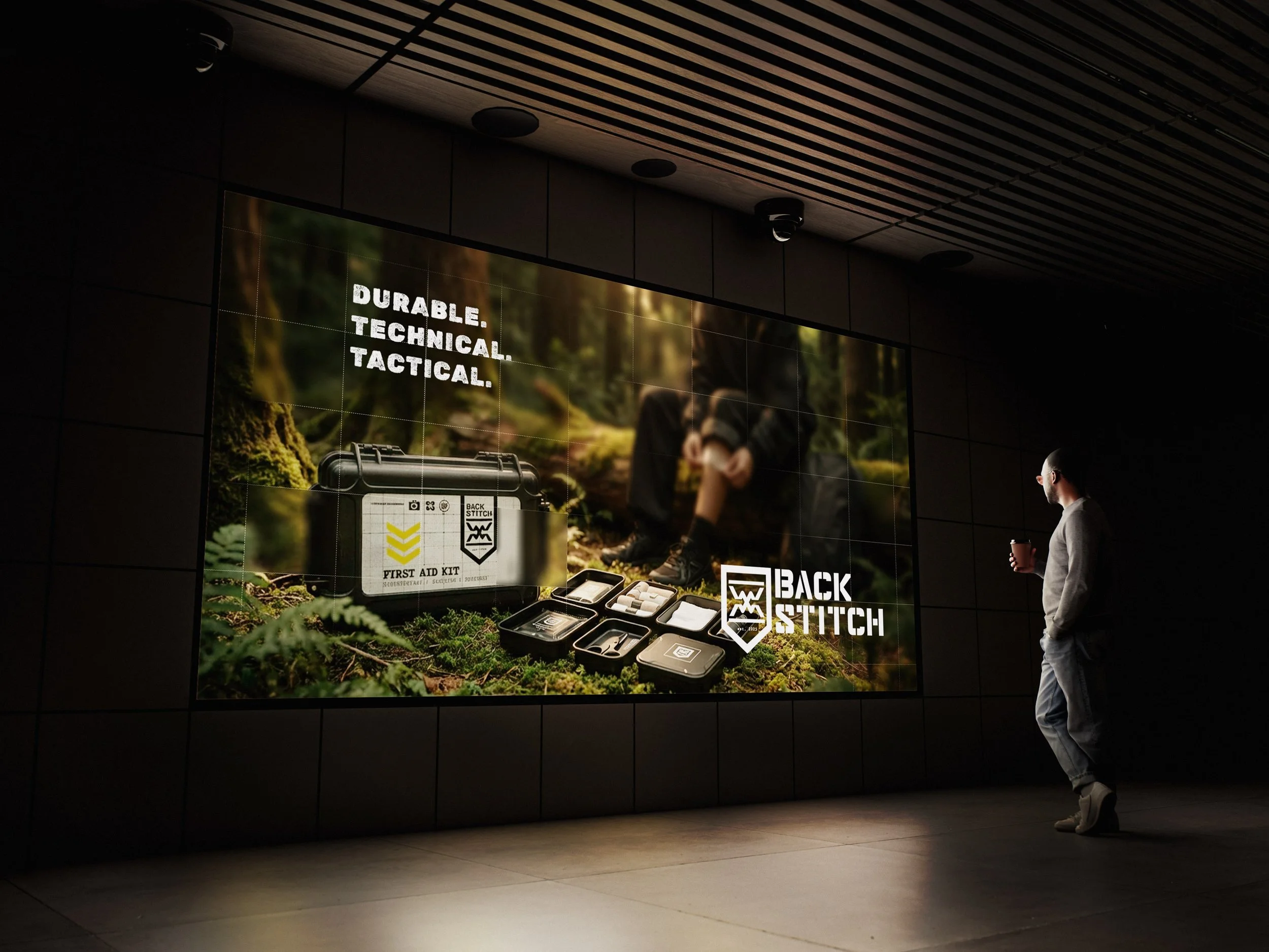 A man standing in front of a large digital advertisement displaying a tactical first aid kit, with the words "Durable. Technical. Tactical." and the brand "Back Stitch".