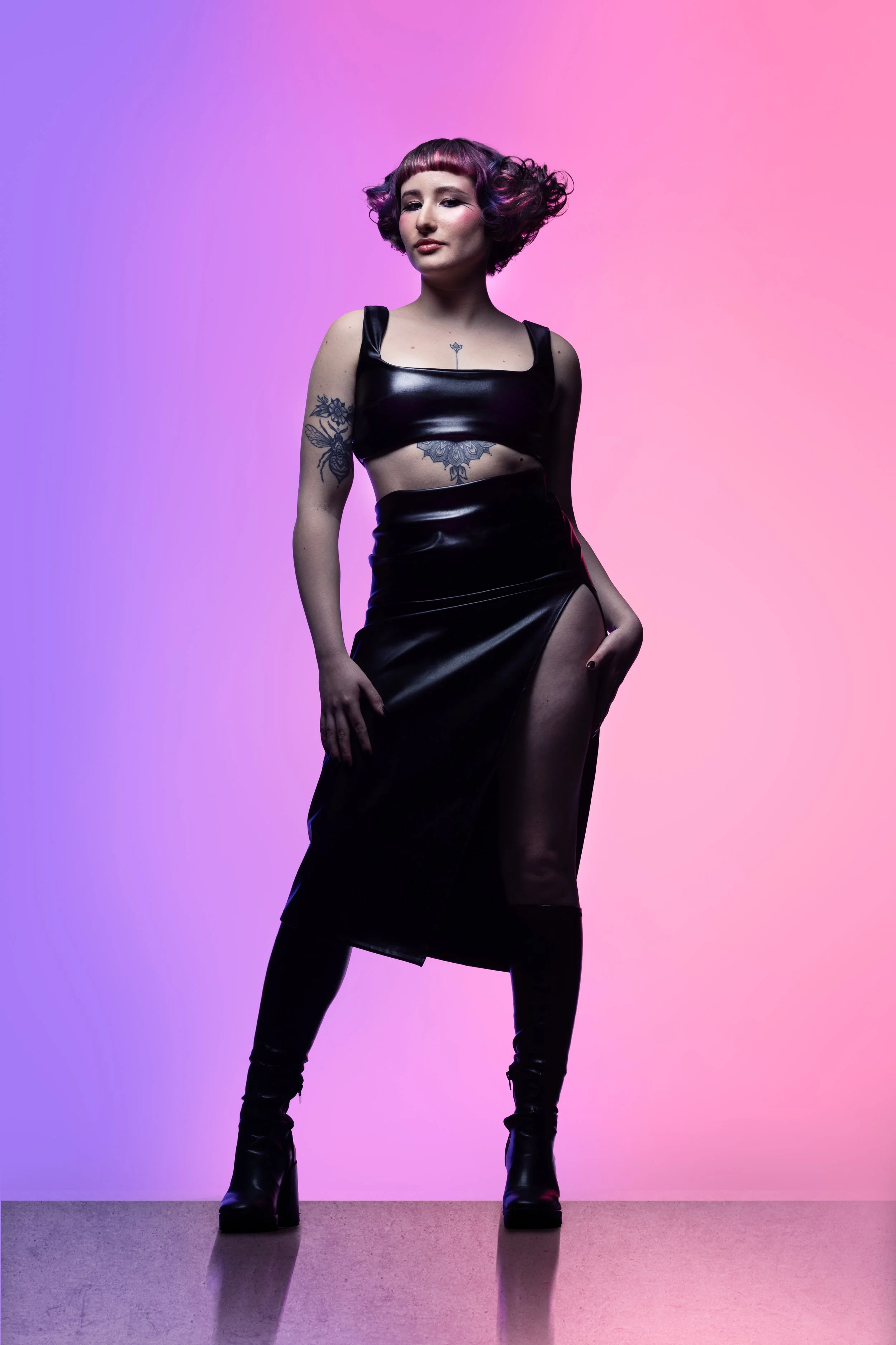 A woman with pink and purple styled hair wearing a black leather dress with a high slit, standing against a colorful gradient background.