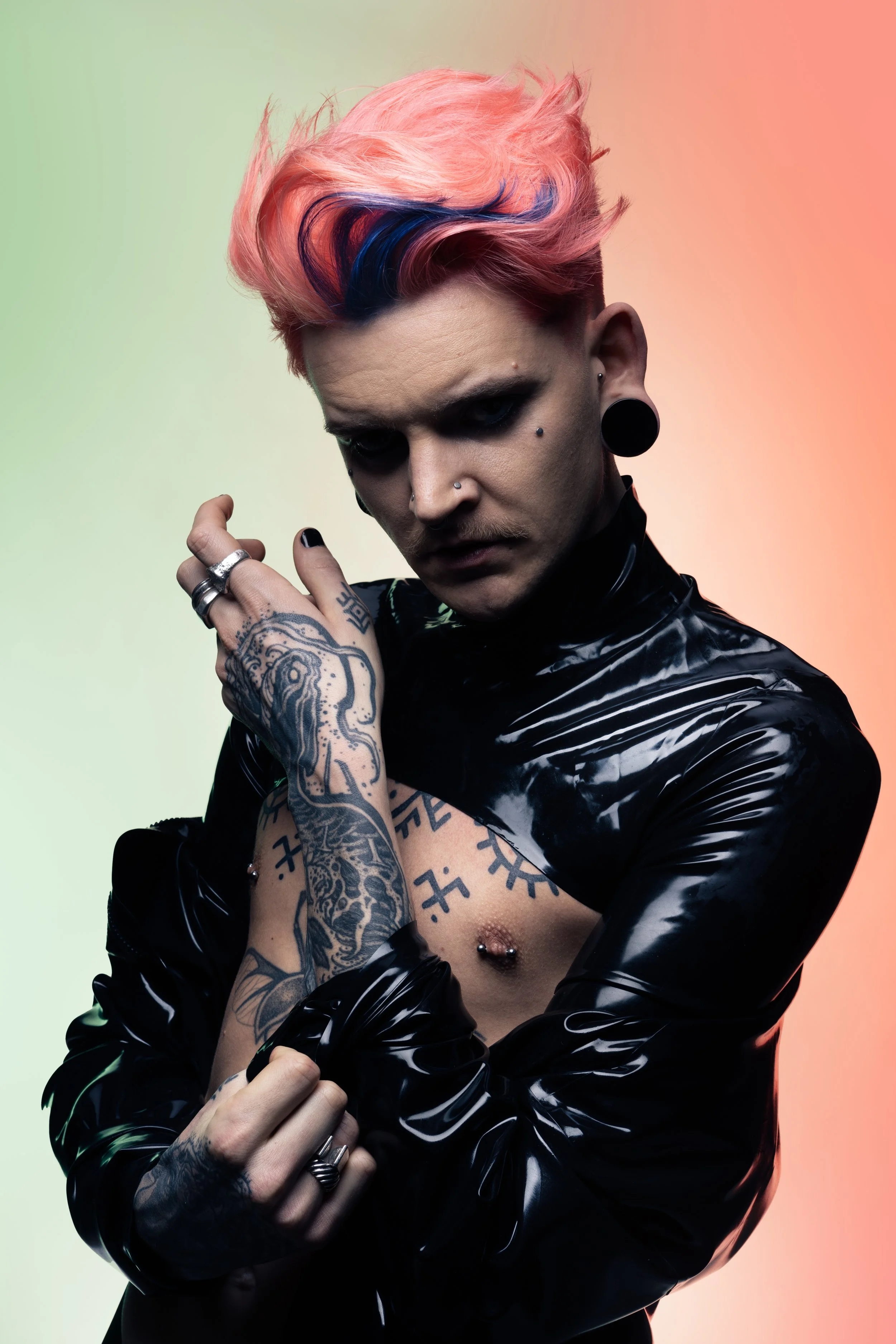 A person with pink, purple, and blue styled hair, wearing makeup, black earrings, and tattoos on arms and chest, posing against a two-tone gradient background.