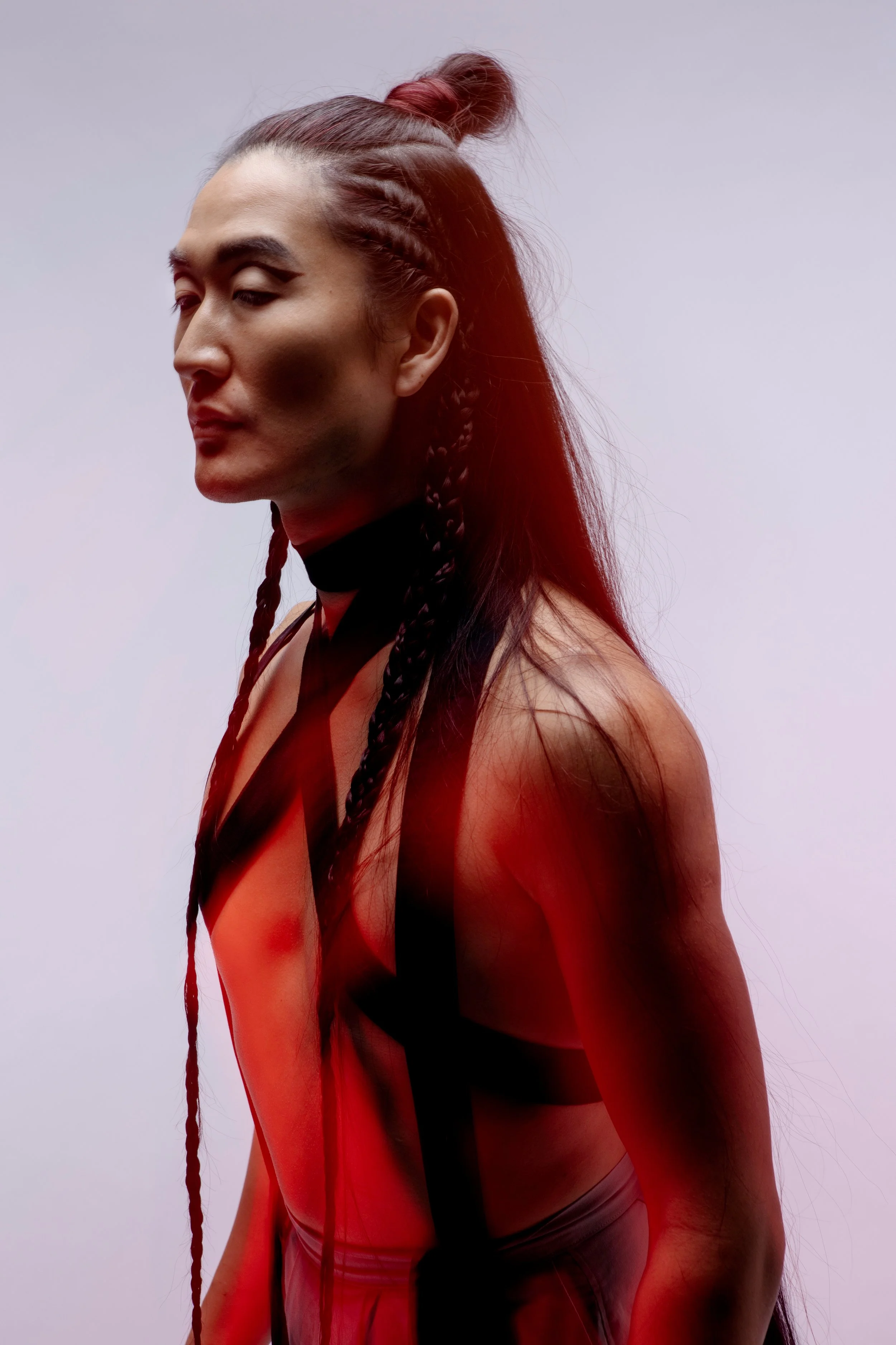 A woman with long, braided hair and a top with black straps, appearing in a fashion or artistic photoshoot with a plain, light-colored background.