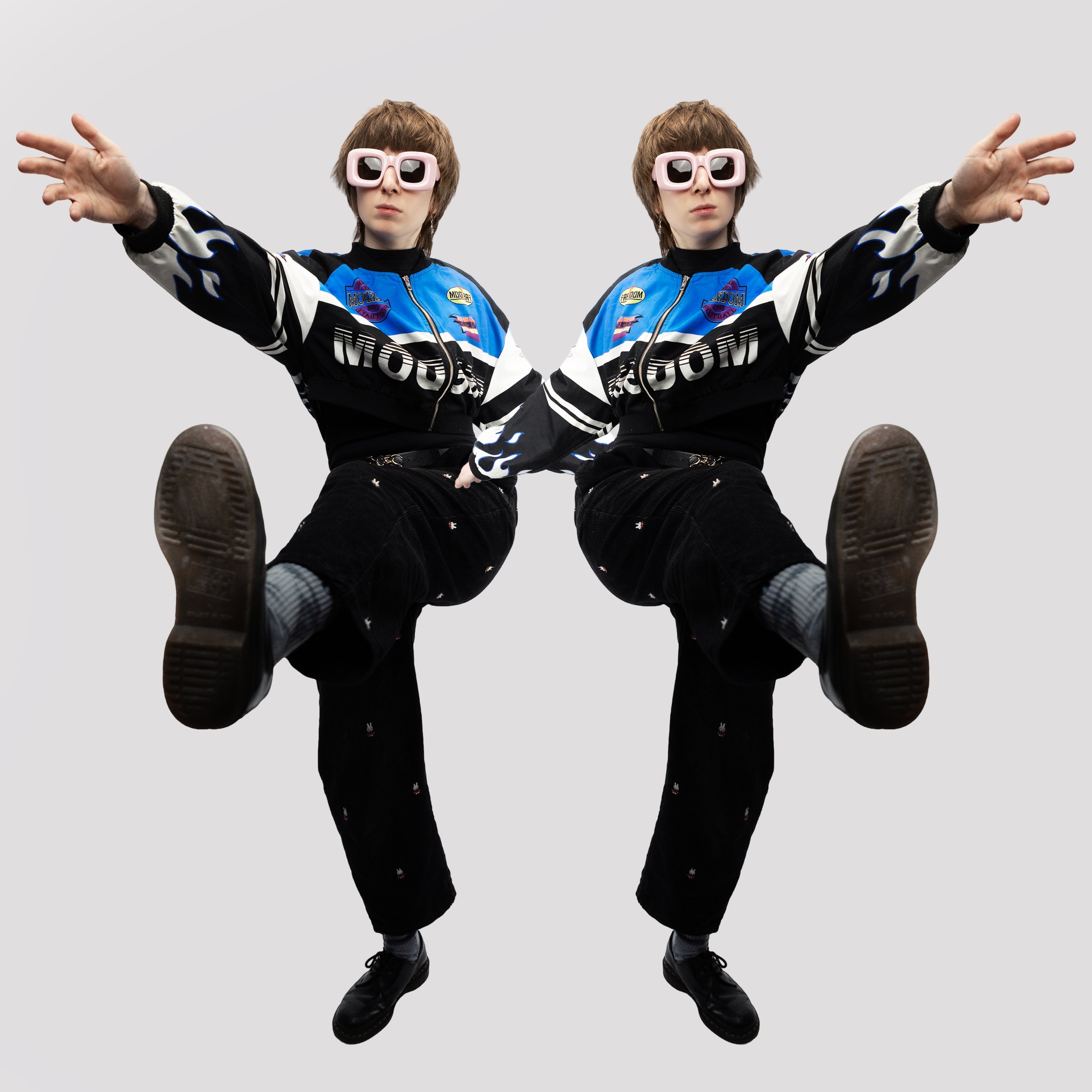 Two identical young men in black and blue jackets, black pants, gray socks, and black shoes, pose with one leg raised and arms extended, wearing large pink sunglasses, against a plain gray background.