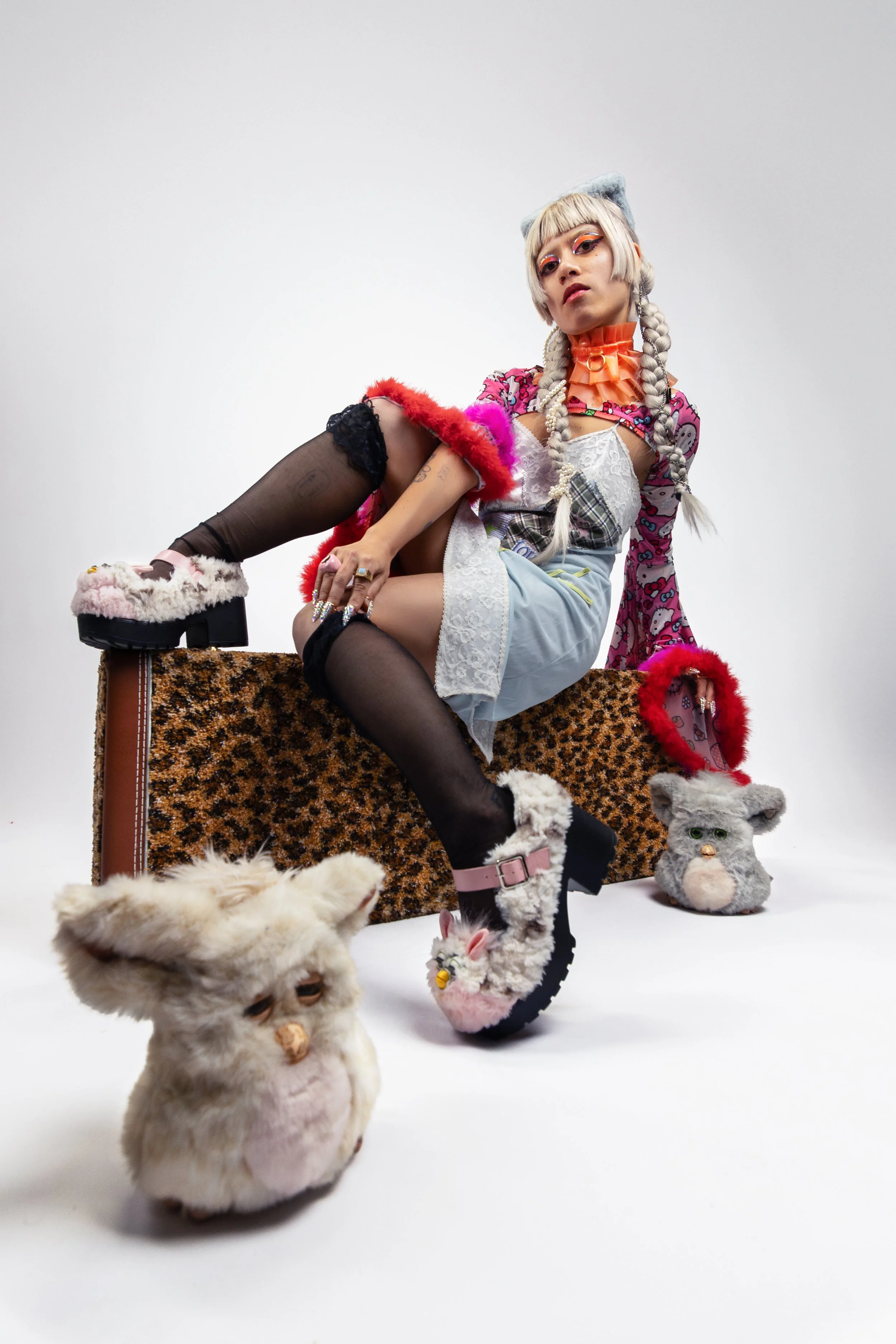 Person sitting on a leopard print bench wearing eclectic fashion including patterned and lace clothing, colorful accessories, and furry shoes, surrounded by plush animal toys, with a plain white background.