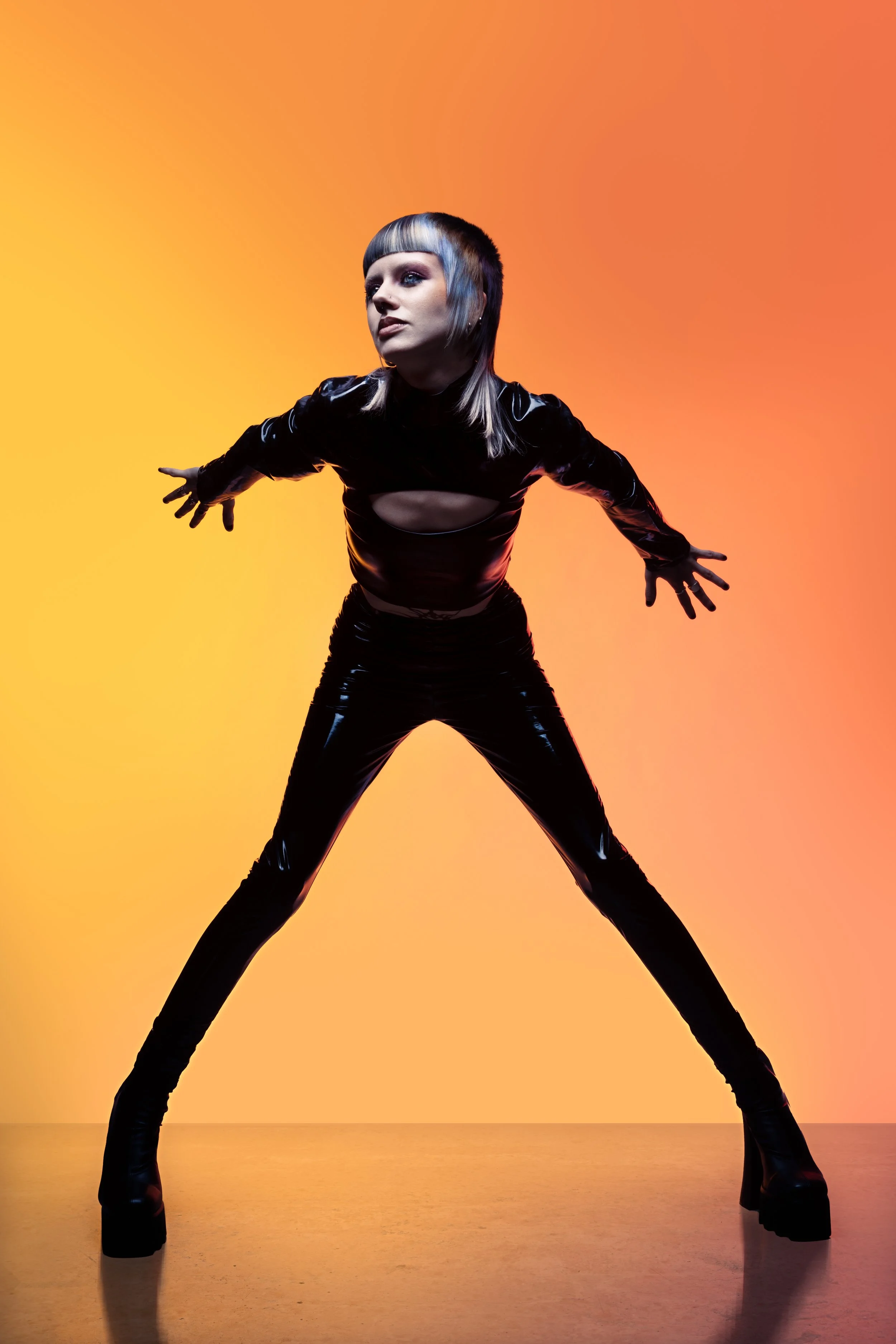 A woman in black latex clothing poses with arms outstretched against a gradient orange and yellow background.