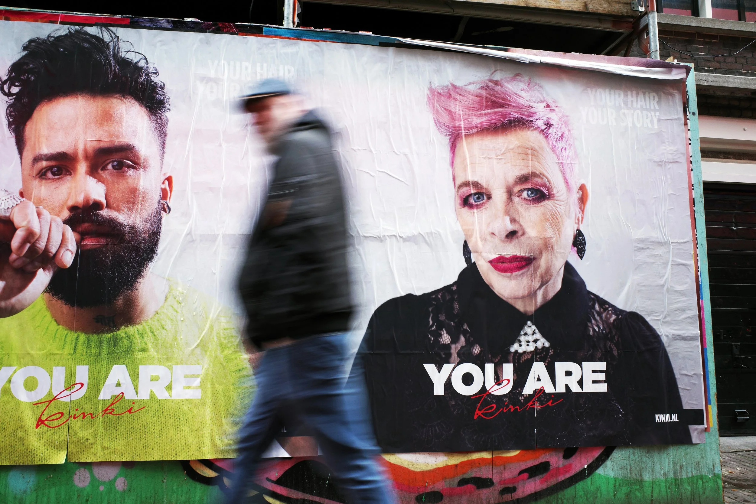 A blurred man walking past a large advertisement billboard showing two women with the text 'YOU ARE' and 'Kink'. The woman on the left has short dark hair and a beard, wearing a bright green top. The woman on the right has short pink hair, makeup, an
