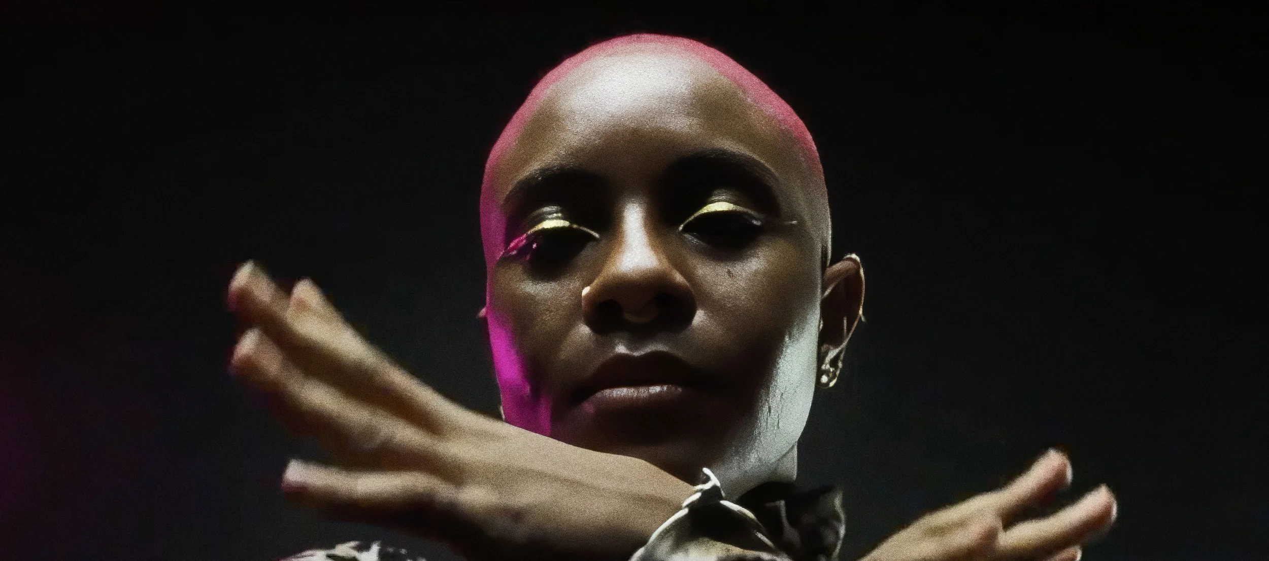 Close-up of a person with short pink hair, gold makeup on eyelids, and an intense expression. They are crossing their arms in front of their face, wearing earrings and a patterned shirt against a dark background.