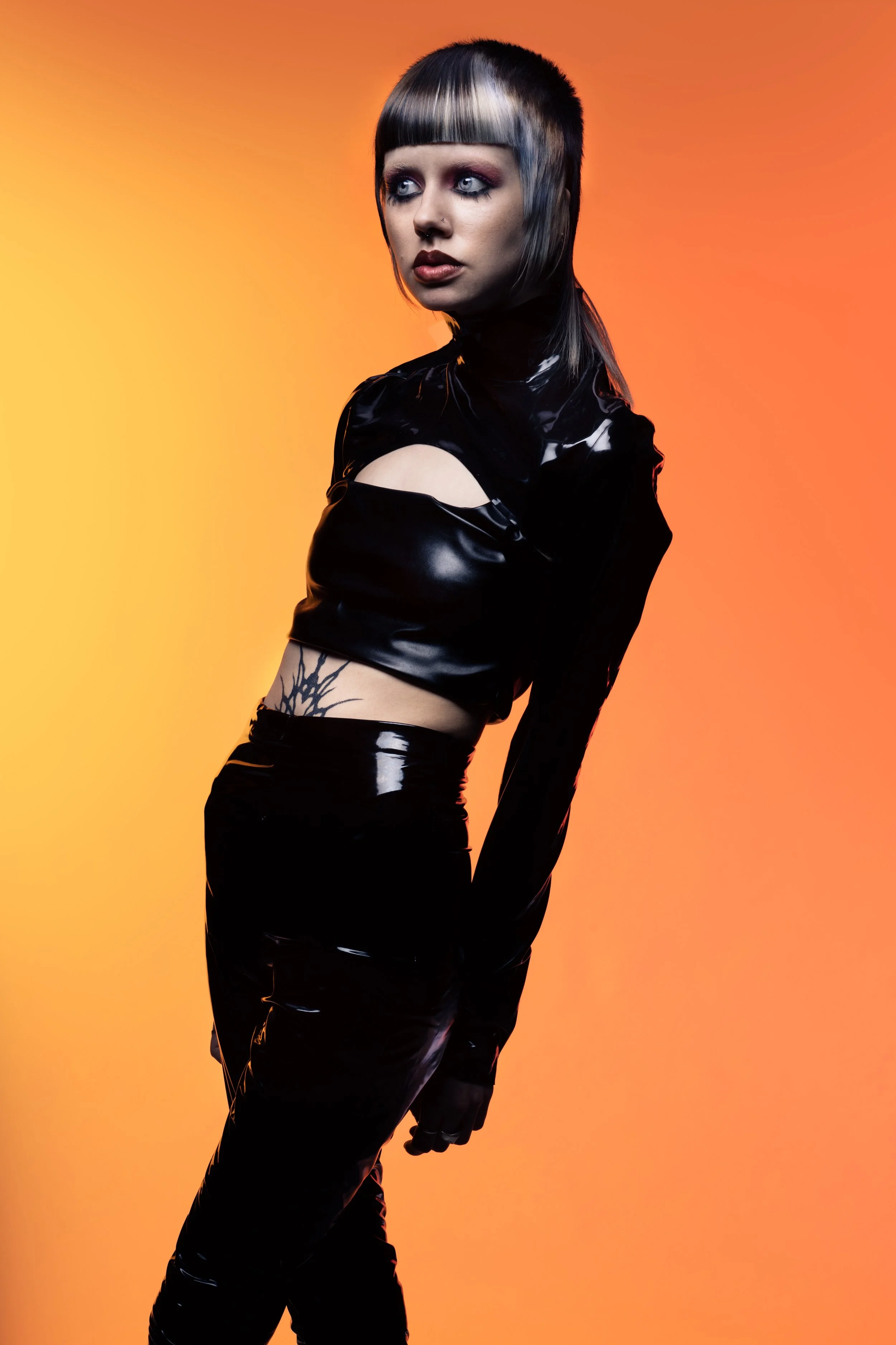 A woman with silver and black hair, wearing black shiny latex clothing, standing against an orange gradient background.
